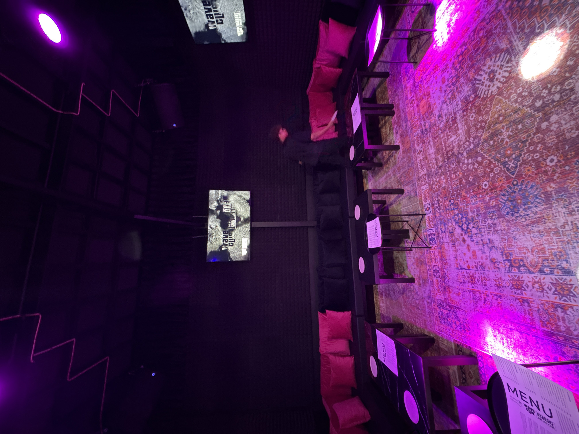 A nighttime interior scene in Baan Bang Rak, Thailand. A carpeted floor is bathed in pink light, leading to a wall with two screens displaying the words "Wave" and "Clip". A person stands between rows of tables and lounge seating, with pink cushions and menu cards on tables. The lighting is predominantly purple.