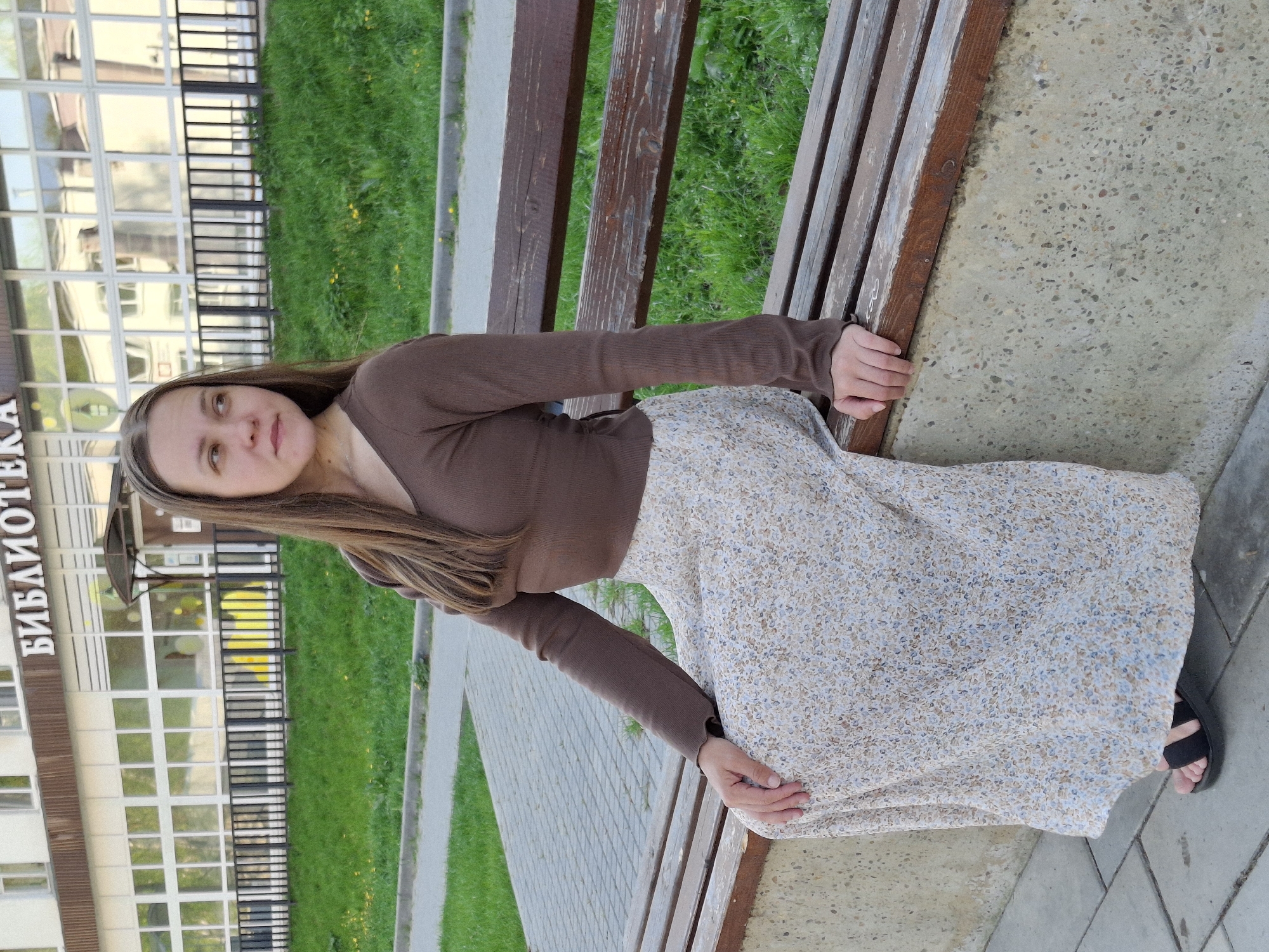 A full-length portrait shows a young woman sitting on a park bench in Chaikovsky, Russia. The subject has long brown hair and is wearing a brown long-sleeve crop top and a cream-colored floral skirt. She is seated on a wooden park bench, facing the camera. To the left of the bench is a green lawn, a sidewalk, and the facade of a multi-story building. The sign on the building reads "БИБЛИОТЕКА" in Russian, indicating it is a library. The surrounding environment suggests an urban park setting.