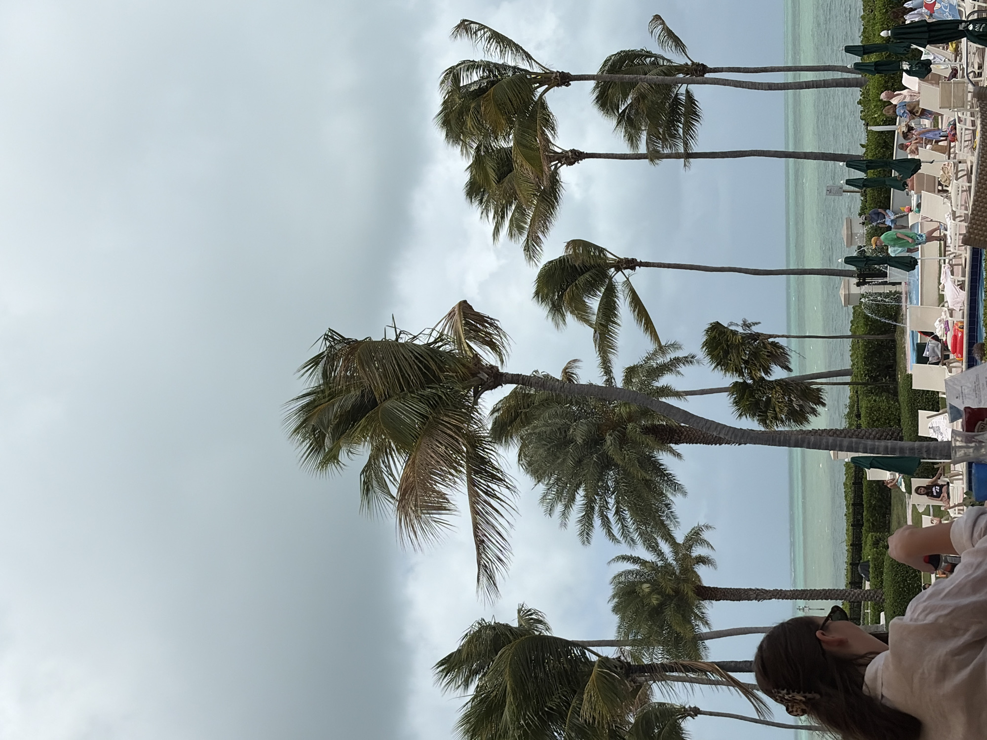 The image shows a scene at what appears to be a resort or hotel in Key Biscayne, United States of America. Palm trees frame the view of the ocean, visible in the background. There's a pool area with lounge chairs and people relaxing or walking around. In the foreground, a person with dark hair and sunglasses is partially visible, suggesting they are seated at a table, possibly at a restaurant or bar, enjoying the view. The sky is overcast, indicating the weather is likely cloudy or potentially rainy. The environment suggests a tropical or subtropical climate characteristic of Key Biscayne.