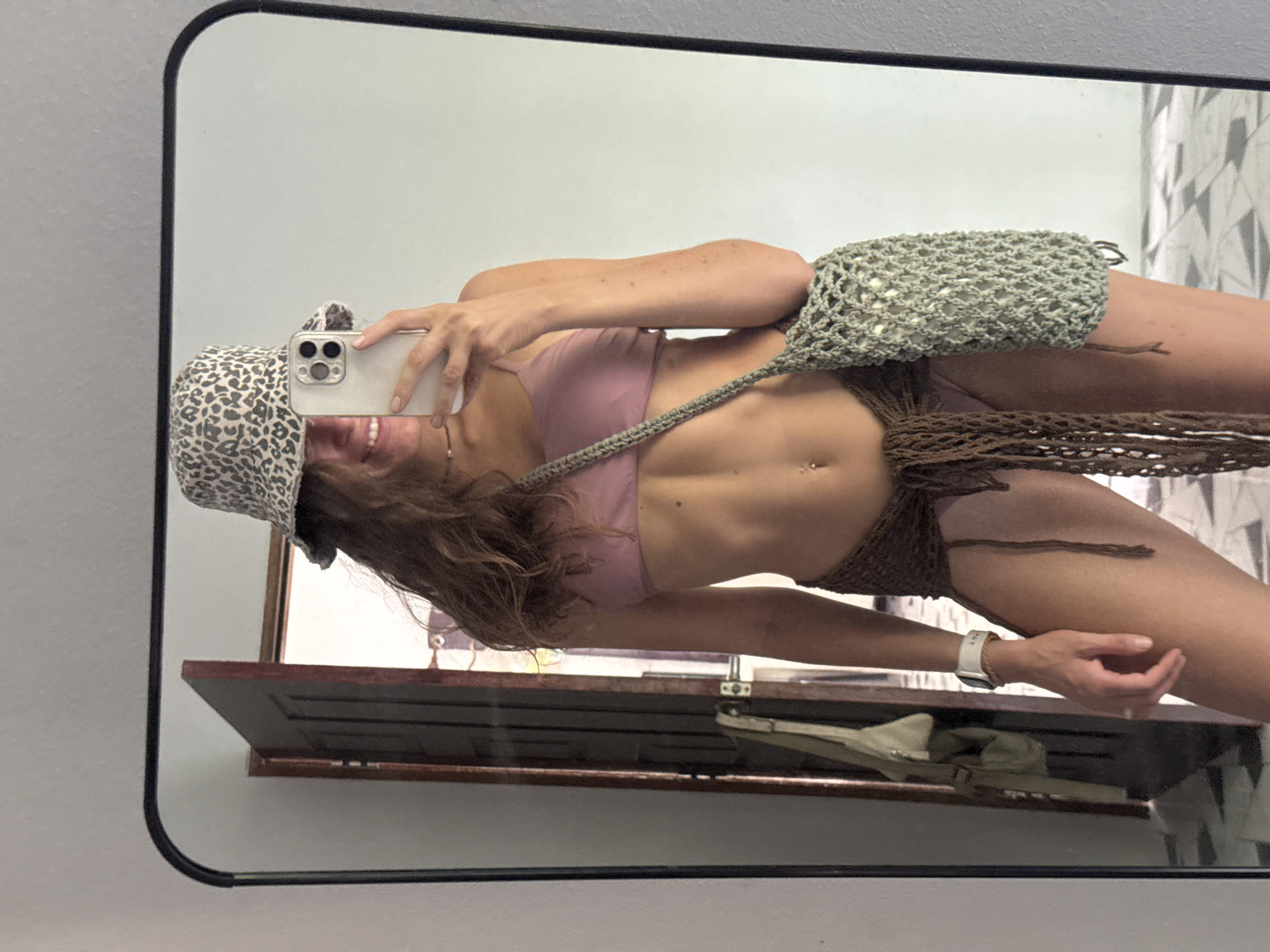 An individual is captured in a mirror selfie, seen from the chest up to the mid-thigh, in what appears to be an indoor setting. The image is rotated 90 degrees clockwise, so the person is horizontally oriented within the frame. They are smiling broadly, showing their teeth, while holding a smartphone with a light-colored case and multiple camera lenses, indicating they are taking the photo themselves.
The person is wearing a leopard print bucket hat, light purple or pink bikini top, and a brown crochet-style skirt or wrap. A light green crochet sling bag is draped over their shoulder and across their torso. They have long, wavy, light brown hair. Notable details include a belly button piercing and a white-banded wristwatch on their left wrist.
The background reflected in the mirror shows a plain light-colored wall to the left and a wall with a geometric pattern, possibly tiles, to the right. A dark brown wooden shelf is visible behind the person, with what looks like a light-colored bag or shoes resting on it. No discernible text is visible in the image. The attire suggests a warm climate or casual, beach-ready fashion.
