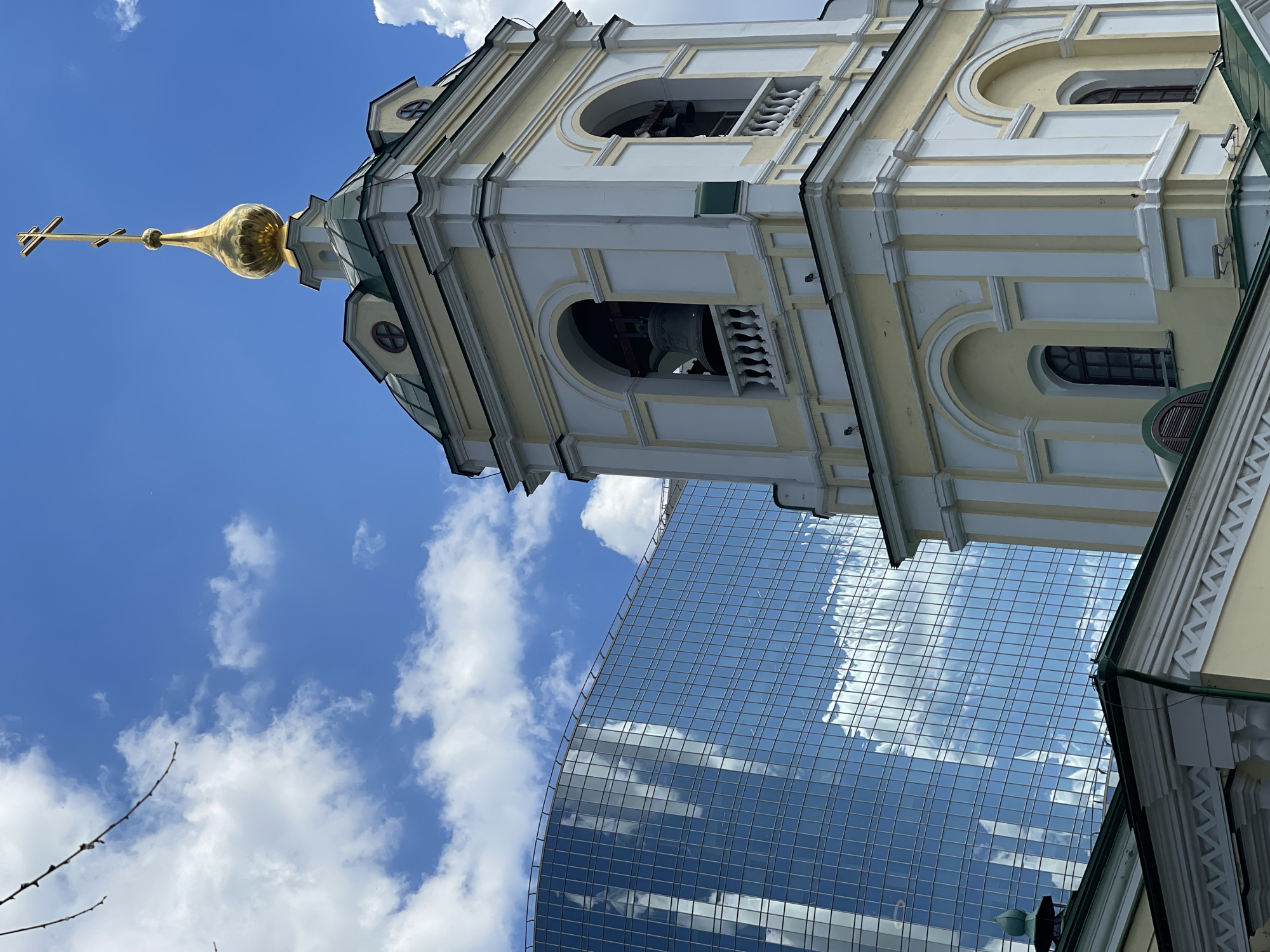 The image presents a striking architectural contrast in Moscow, Russia, featuring a historic Orthodox church bell tower alongside a modern glass skyscraper. The bell tower, rendered in light yellow and white, showcases classical architectural elements with multiple tiers of arched openings, ornate pilasters, and decorative balustrades. Large bells are visible within the open arches of its upper sections. The tower is crowned with a golden, fluted onion-shaped dome, from which a prominent golden Orthodox cross extends upwards, gleaming in the sunlight.
Adjacent to and partially behind the bell tower, a contemporary skyscraper rises, its entire facade clad in reflective glass panels. These surfaces brilliantly mirror the vibrant blue sky and scattered fluffy white cumulus clouds, creating a dynamic visual effect. The sleek, grid-patterned glass of the modern building offers a stark visual and stylistic contrast to the intricate and traditional design of the religious structure.
The photograph is taken from a low-angle perspective, looking upwards, which emphasizes the towering presence of both edifices against a clear, bright blue sky indicative of a sunny day. This juxtaposition of an ancient religious monument and a futuristic high-rise vividly illustrates the blend of historical heritage and modern urban development characteristic of the city. There are no people or visible text in the scene.