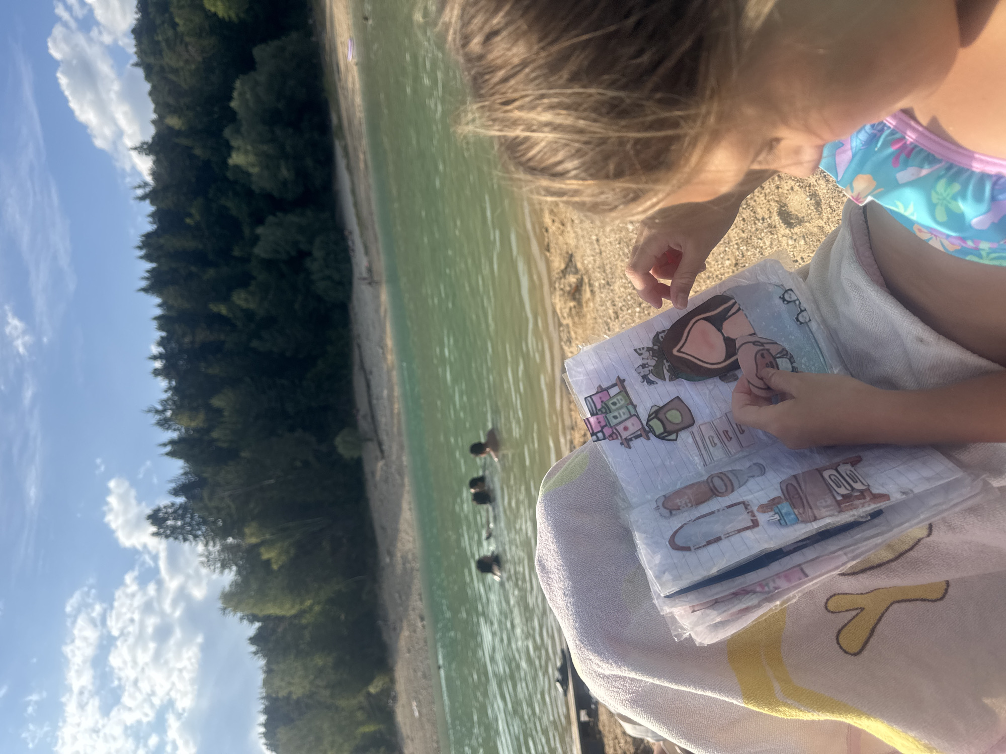 In Maisod, France, a young girl with blonde hair is sitting on a beach, holding a booklet with paper cutouts of clothing and accessories. The booklet is open, and she is looking at the cutouts. The girl is wearing a blue and pink floral swimsuit. In the background, a lake is visible with several people swimming in it. A tree-covered hill borders the lake. The sky is blue with scattered white clouds. The image is taken from a close-up perspective, with the girl's back to the viewer, focusing on her and the booklet.