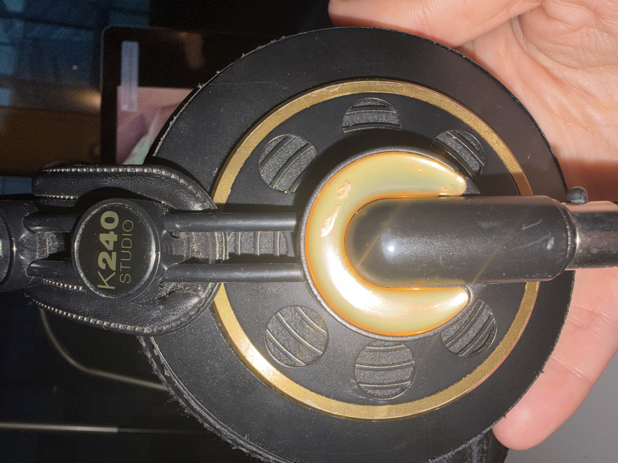 A close-up shows one earcup of an AKG K240 Studio headphone. The earcup is circular and primarily black, with a thin gold ring around the center. A cream-colored, crescent-shaped element is in the middle. The model number "K240 STUDIO" is visible on the black plastic connecting piece. A human hand is visible, partially covering the right side of the earcup. The location is unknown, as no information is provided about the specific location in L'Hospitalet De Llobregat, Spain or any other location.