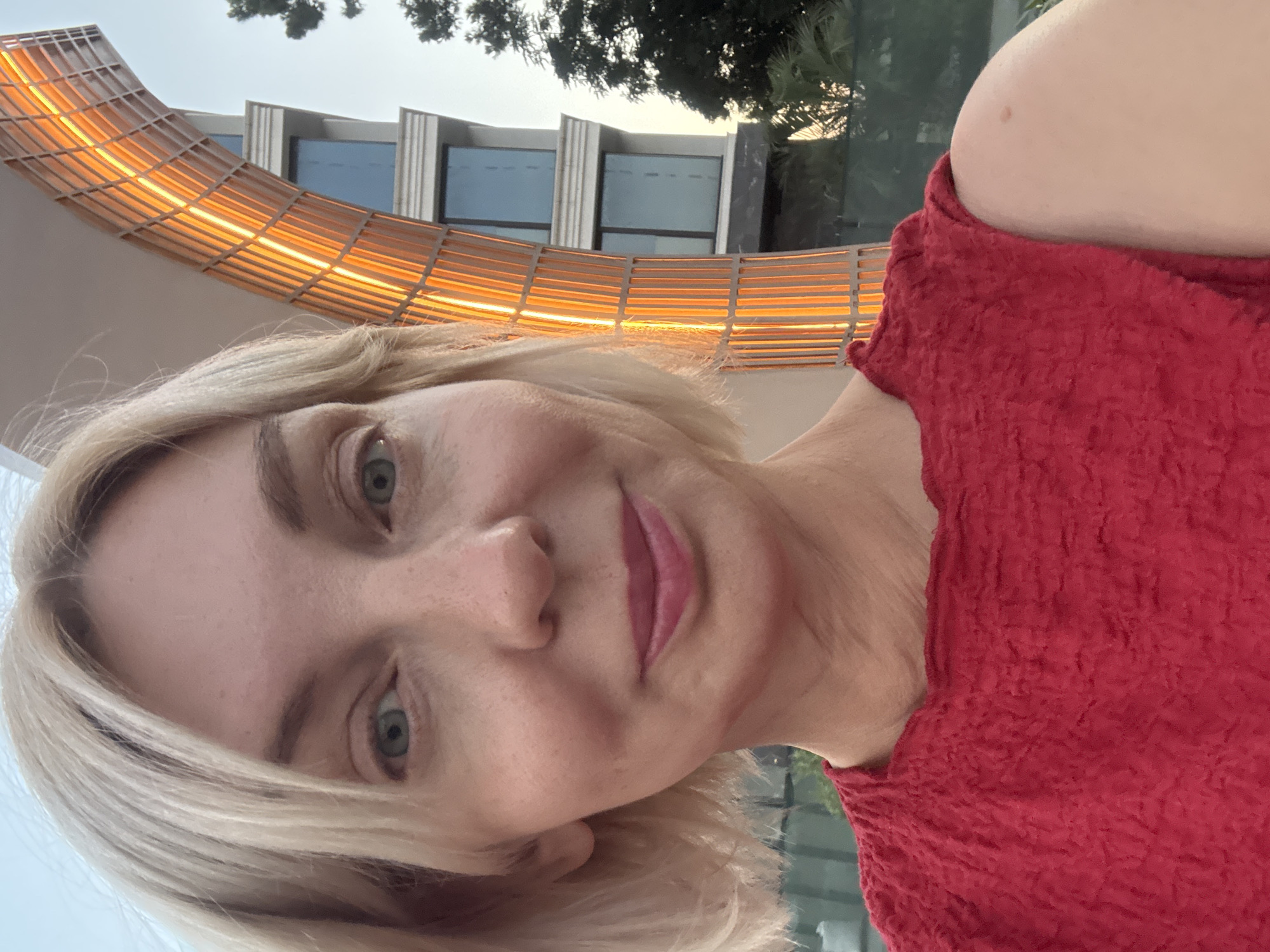A woman with blonde hair and blue eyes is taking a selfie outdoors. She is wearing a red top. Behind her, an illuminated structure with a curved design is visible, along with a modern building and some trees. The lighting suggests it is either early morning or late evening. The location is identified as Sochi, Russia.