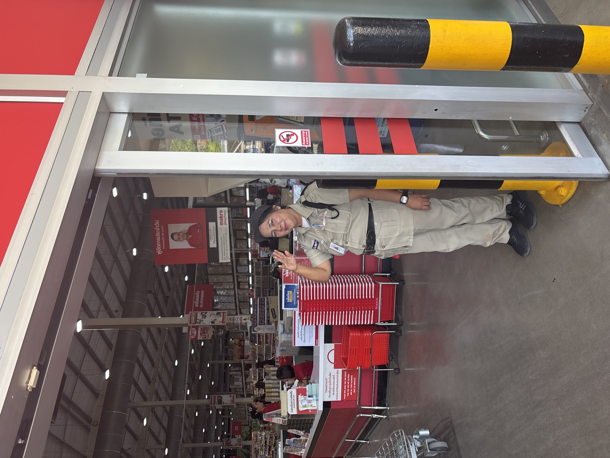 A full shot captures a security guard at the entrance of a Makro store in Baan Bo Phut, Thailand. The guard, wearing a beige uniform and a black cap, stands near red shopping baskets, waving with a friendly gesture. The store's interior is visible, showing shelves stocked with products. A sign near the entrance prohibits firearms. The building's framework includes red panels and a black and yellow striped barrier.