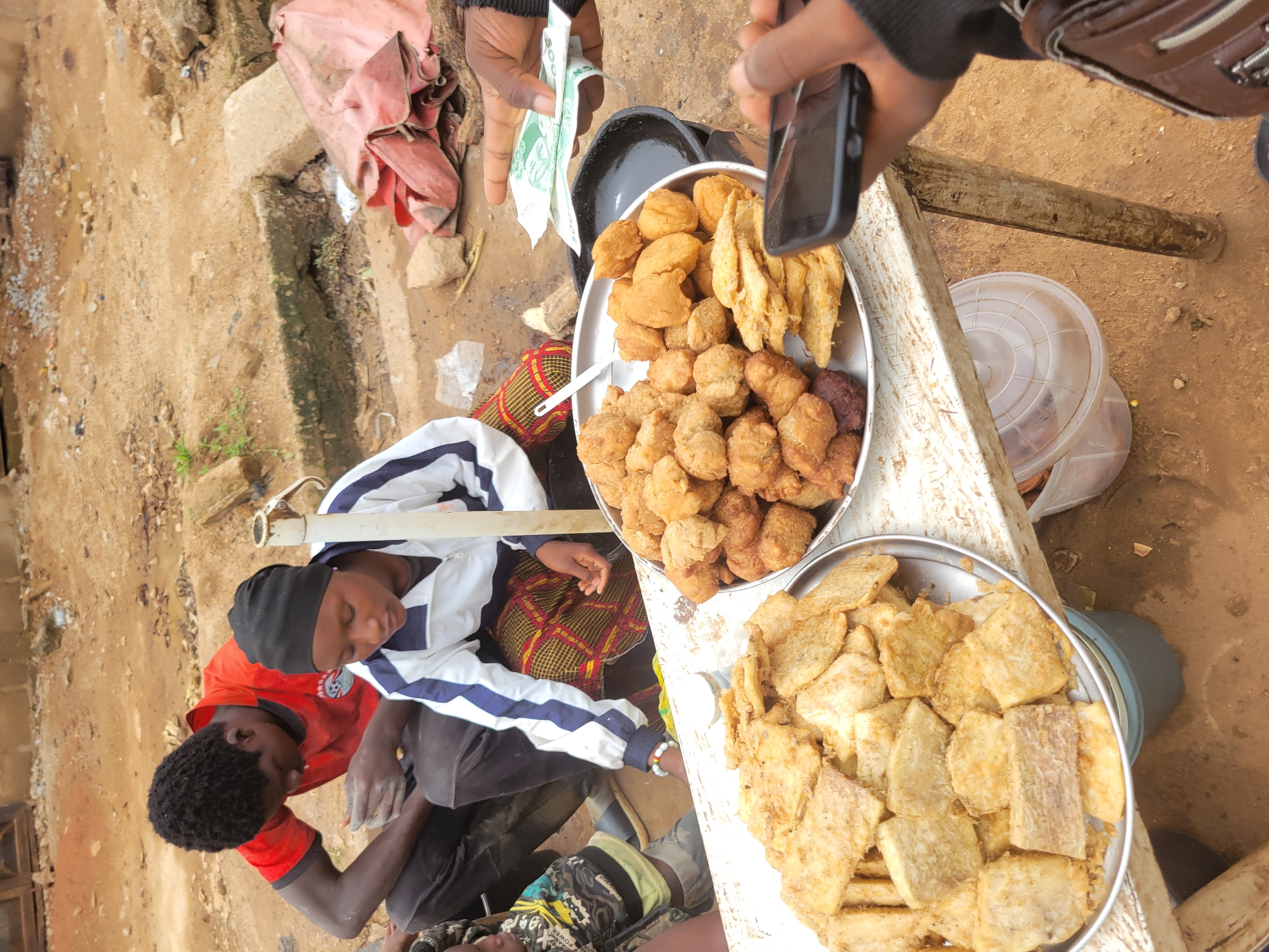 In Bokkos, Nigeria, a street vendor displays an assortment of fried snacks. Two individuals are visible in the foreground: a man in a red shirt and a woman wearing a black headscarf and a white and blue jacket. They appear to be engaged in a transaction with a customer whose hand is holding money and a smartphone.
The snacks are arranged on two metal platters on a weathered wooden table. The platter on the left contains slices of what appears to be fried plantain or yam. The platter on the right holds a mixture of round, golden-brown fried items and some flatter, fried pieces. The background shows a dirt ground with some debris and a pink tarp. The scene suggests a bustling marketplace or roadside stall, typical of urban or semi-urban Nigerian settings.