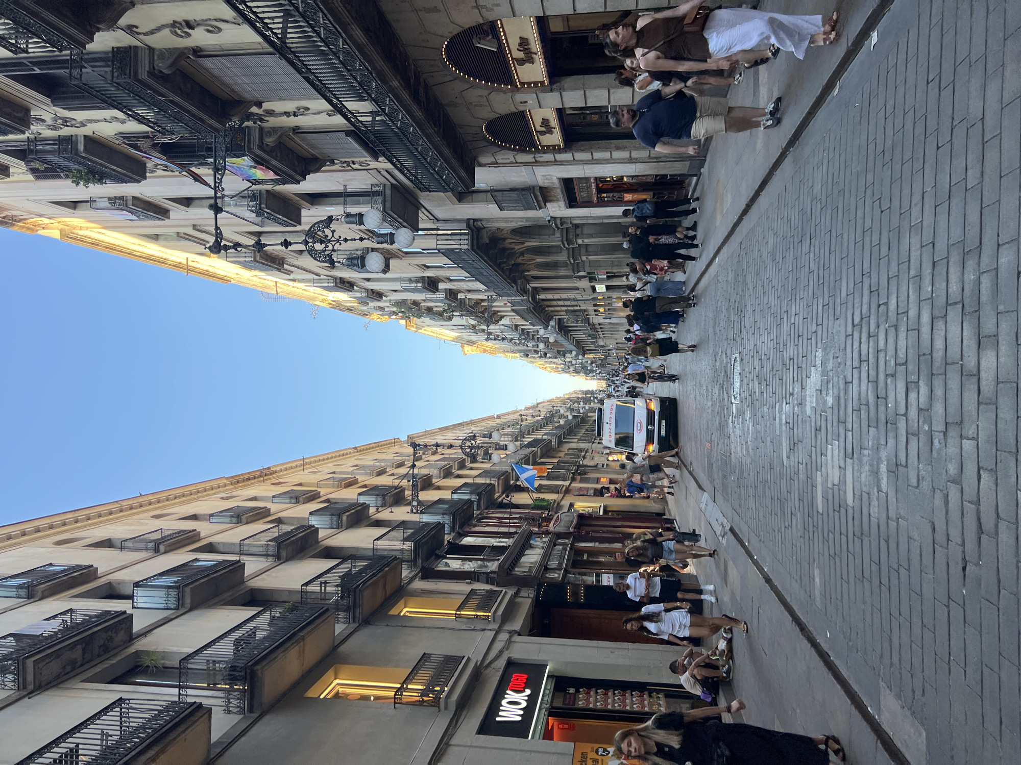 The image depicts a narrow, pedestrianized street lined with tall, multi-story buildings on both sides. The architecture suggests a European city, with wrought-iron balconies adorning the upper floors of the buildings. The street is paved with cobblestones and is filled with people walking in both directions. A white van is visible in the distance, navigating the street. Shops are present on the ground floor of the buildings, with signage visible, including "WOK TO GO" on the left side. The sky is clear and blue, indicating daytime. The scene conveys a lively urban atmosphere, typical of a city center. The presence of flags and the style of the buildings are consistent with Barcelona, Spain, particularly in an older part of the city.