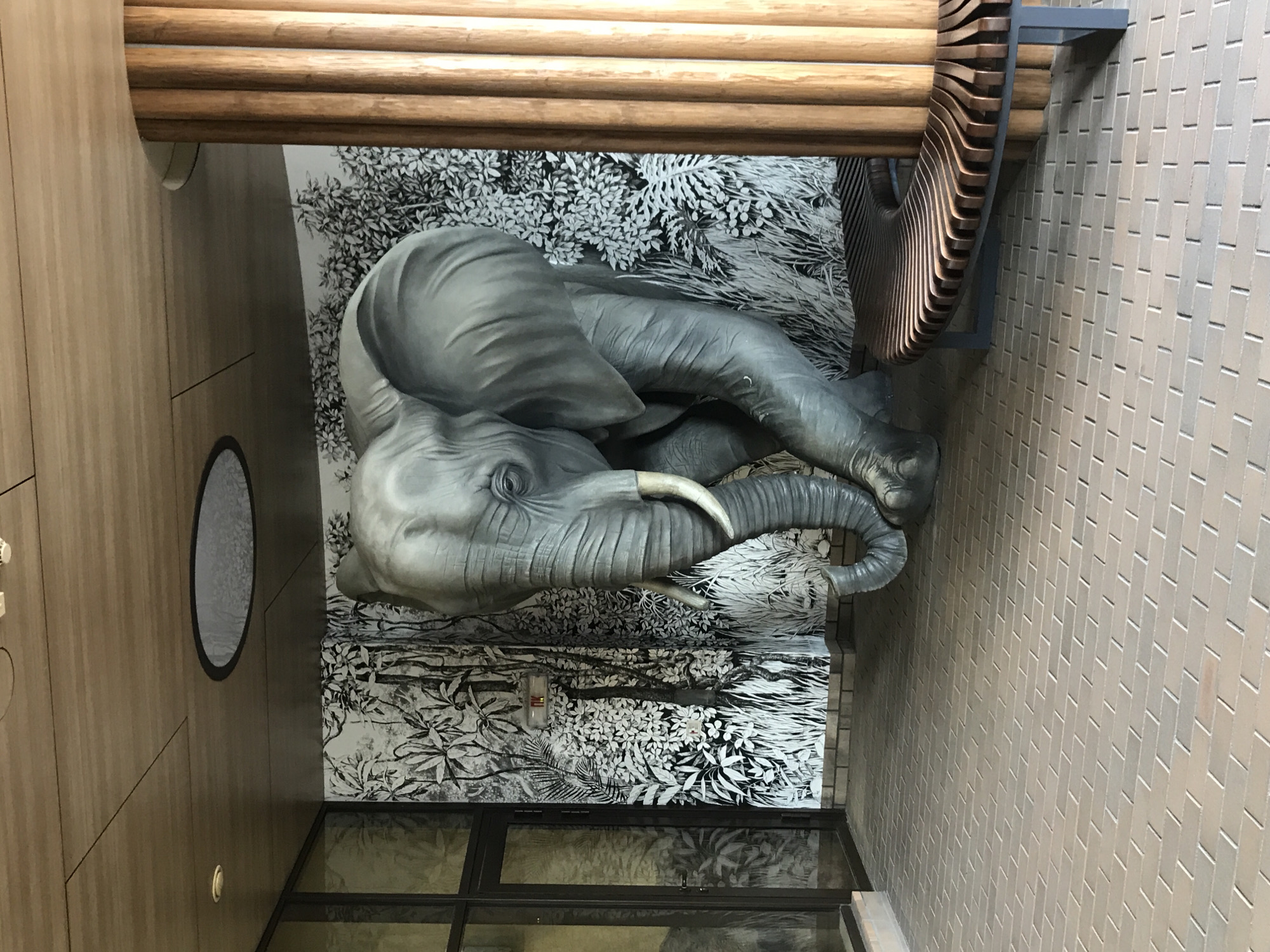 The image shows a large, gray elephant sculpture positioned in front of a black and white mural depicting a jungle scene. The sculpture is detailed, with wrinkles and textured skin. It is walking towards the viewer, with its trunk curved downwards. To the right of the elephant, there is a curved wooden bench with metal legs, placed against a wooden pillar. The floor is paved with light brown bricks laid in a staggered pattern. In the background, a glass door and window are visible. The ceiling is made of wooden panels with a circular light fixture. The scene appears to be an indoor public space, possibly a plaza or a covered walkway. The location is identified as Kharkiv, Ukraine, and the image was taken outdoors.