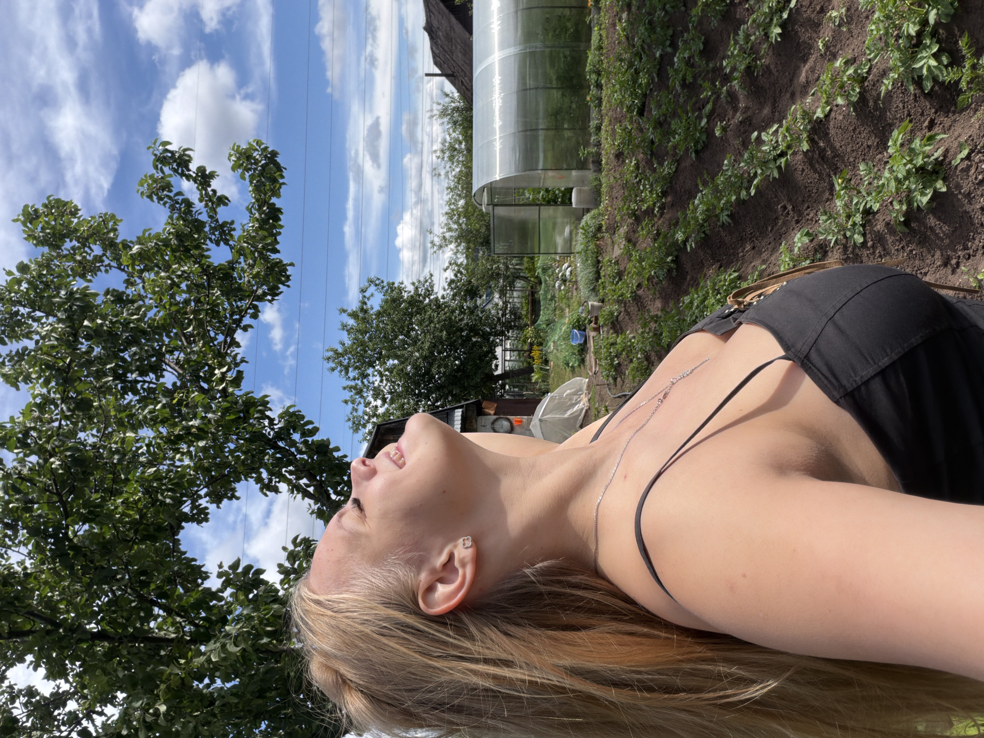 In a garden, a young woman with blonde hair, wearing a black tank top, is looking up with her eyes closed and a smile on her face. Her head is tilted back and her hair is blowing in the wind. In the background, there is a greenhouse, a tree, and a garden bed with plants. The sky is blue with white clouds. This scene is consistent with a dacha, a common type of country house or cottage found in Russia, including in the city of Chaikovsky.