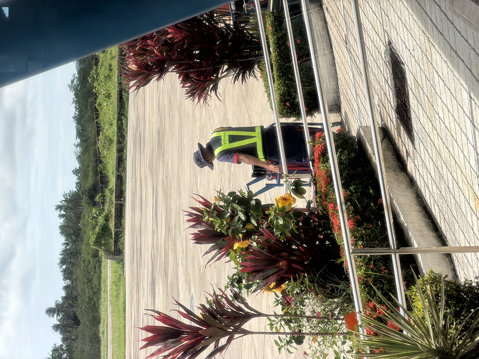 The image shows a man wearing a safety vest and hat, bent over near plants and a railing. He appears to be tending to the vegetation. The setting appears to be a paved area alongside a building with a brick-like exterior, which is in Del Carmen, Philippines. In the background is a green, tree-lined area under a partially cloudy sky. The image is taken at an angle, partially obstructed by an upper frame.