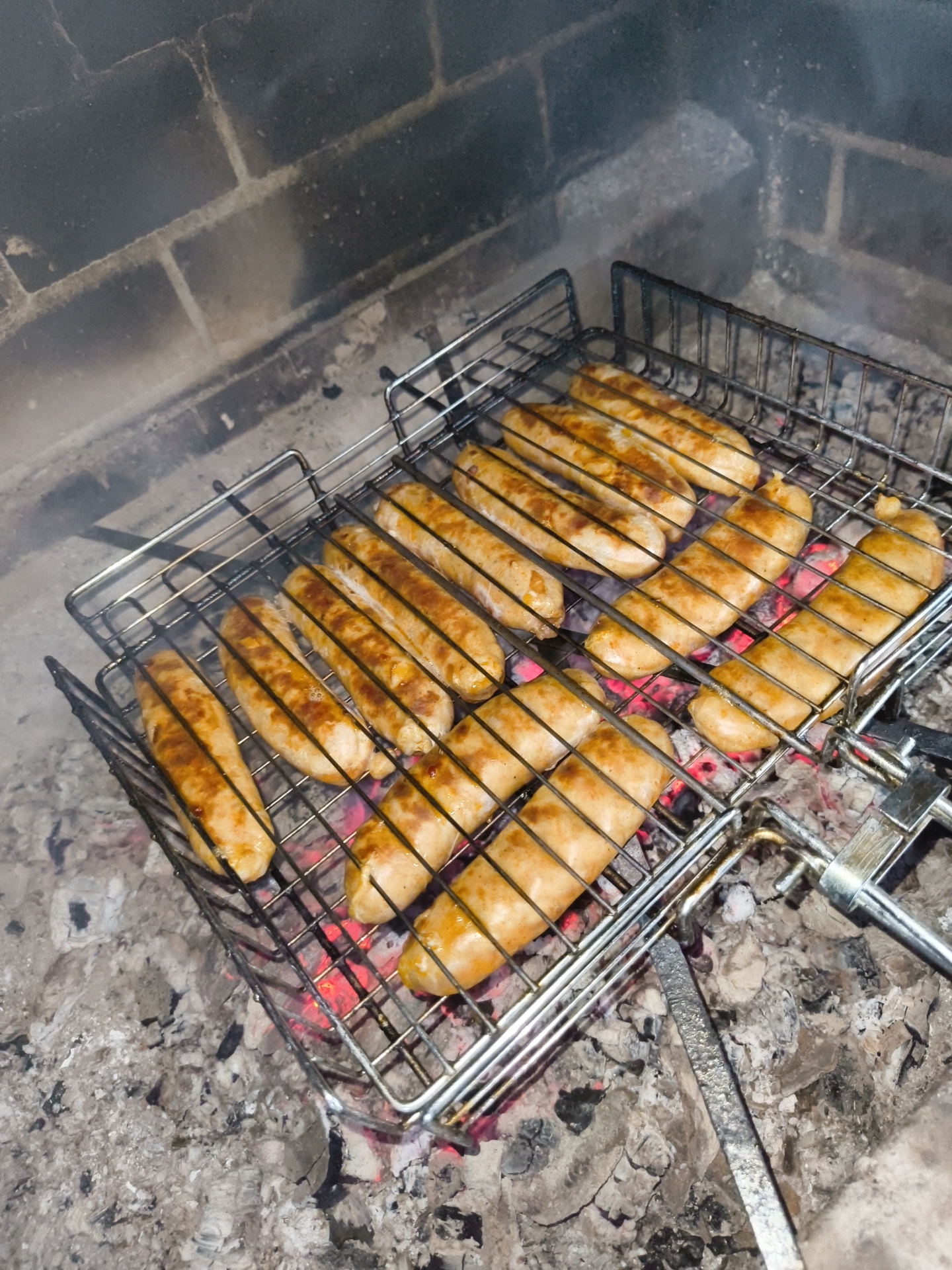A metal grill basket filled with browned sausages is positioned over hot coals in a brick fireplace. The sausages are arranged in rows, and some appear to be slightly charred. Smoke is rising from the grill and the coals glow with a reddish-orange hue. The background consists of dark bricks, typical of a fireplace. The scene appears to be outdoors, possibly in the city of Vostets SNT, Russia, given the context. There are no people visible in the image.