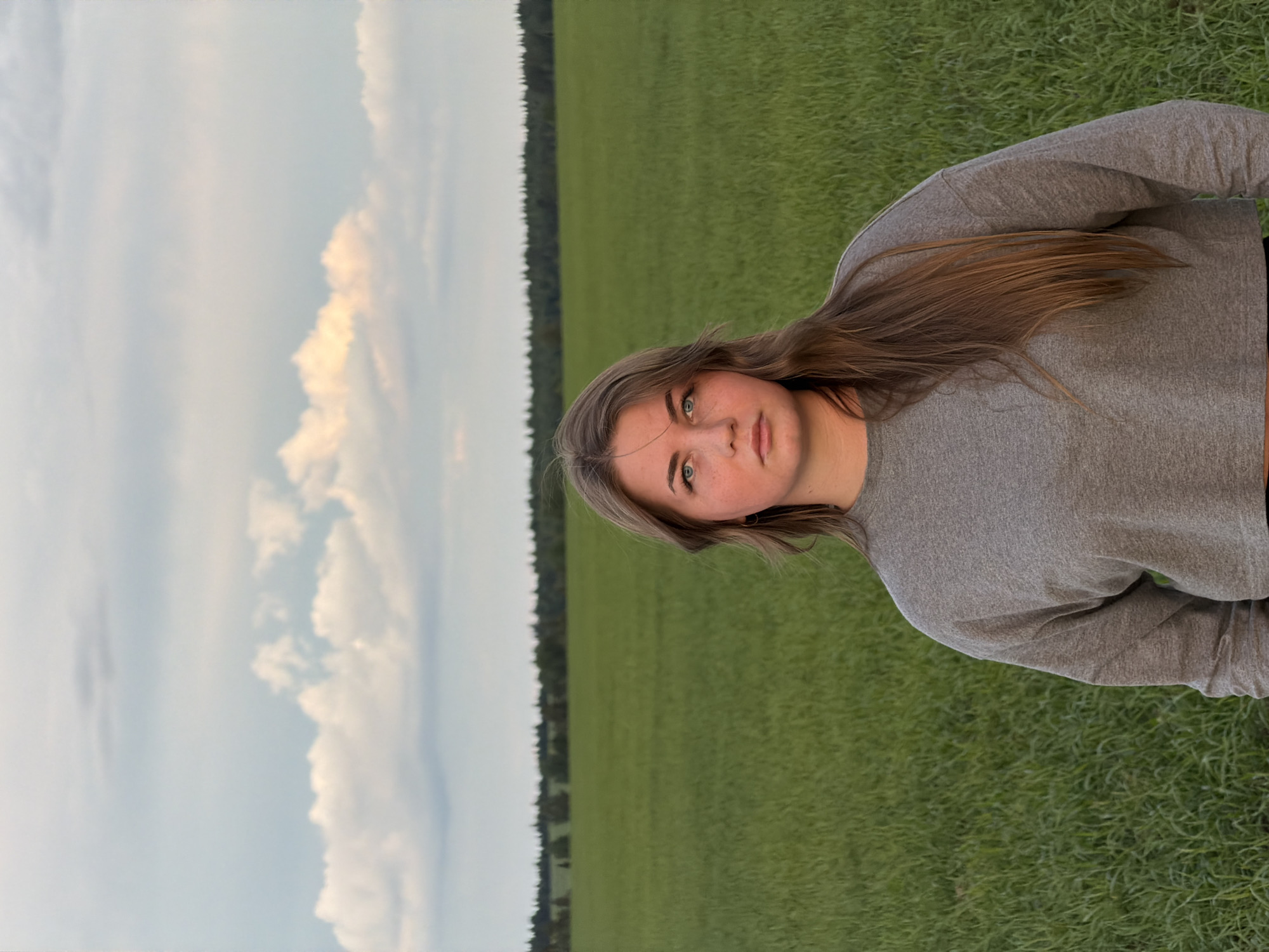 A medium-shot photograph captures a fair-skinned female, likely in her late teens or early twenties, against a landscape of expansive green grass and a partly cloudy sky. She wears a gray long-sleeved shirt. In the background are a strip of dark green trees, likely a forest, along the horizon. The location is not possible to determine based on visual elements in the image, and Фоки, Russia, cannot be confirmed.