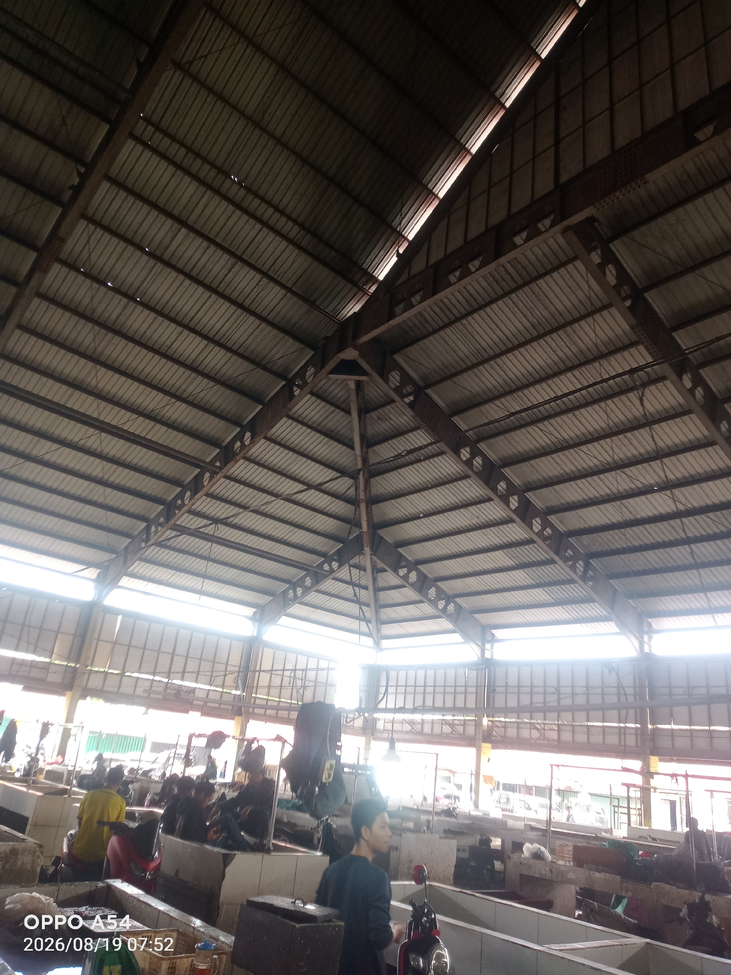 This image captures the interior of a covered market, likely a wet market, in Jambi, Indonesia. The overhead view prominently displays a large, angular corrugated metal roof supported by a complex network of dark metal beams and trusses, forming a distinctive octagonal peak at the center. Natural light streams in from the sides through translucent panels integrated into the roof structure, illuminating the market stalls below.
The market itself appears to be a bustling commercial space with multiple vendor stalls. In the foreground, several individuals are visible. One person in a yellow shirt is seated, facing away from the camera. Further back, more people are engaged in activities at the stalls, some appearing to be vendors and others customers. Various items are present at the stalls, including what seem to be produce or other goods, along with some bags and containers. A red scooter is parked in the lower center of the image. The overall environment suggests a lively, functional marketplace, characteristic of urban settings in Indonesia. The timestamp in the lower left corner indicates the image was taken on August 19, 2026, at 07:52.