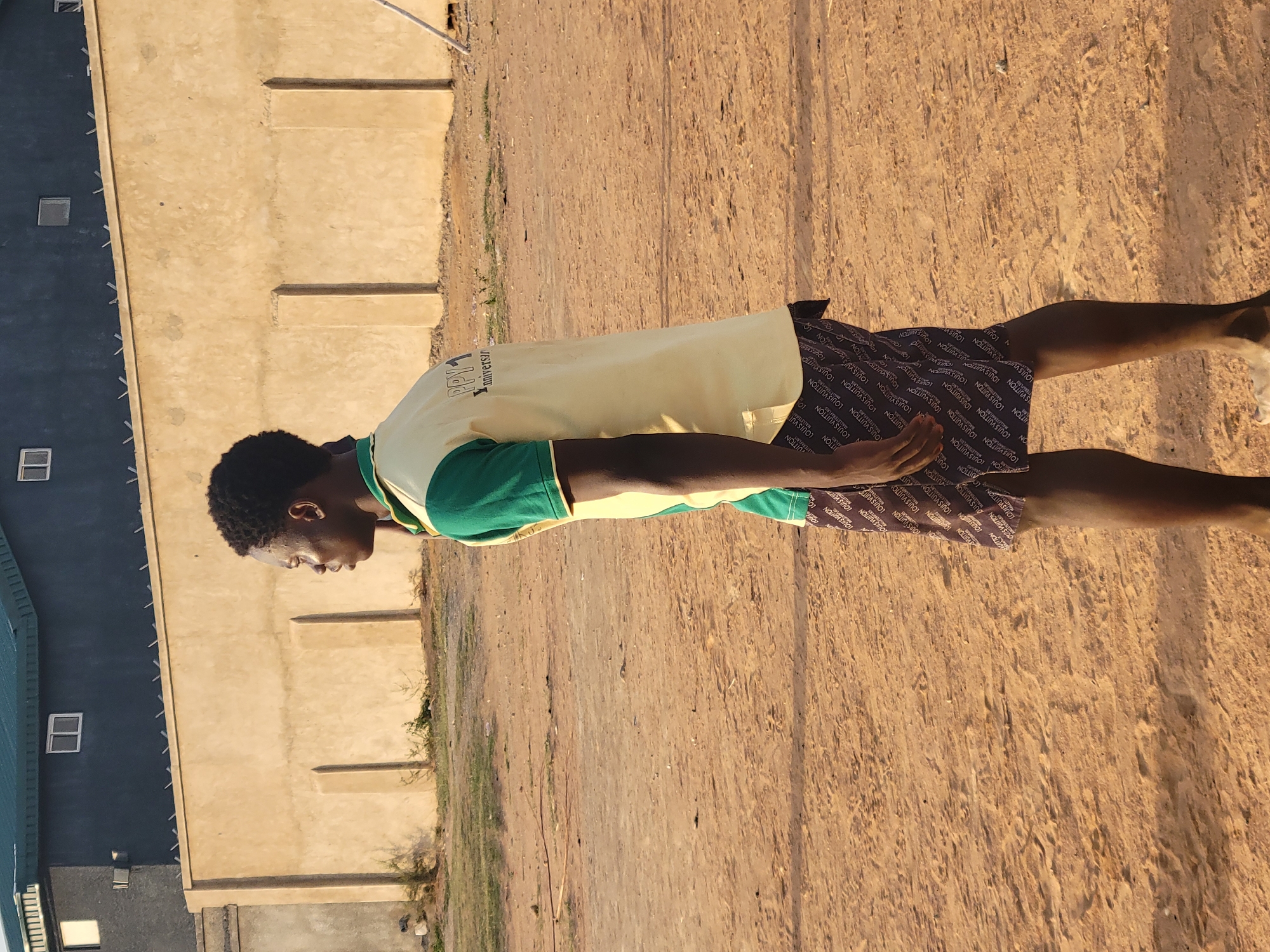 A young Black man stands on a dirt ground in Ibo, Nigeria. The man wears a two-toned shirt with a light yellow body and green sleeves, and dark shorts covered in the Louis Vuitton logo. He is facing the left of the frame, standing perpendicular to a beige building with regular wall indentations. Behind the building and to its left is a building with a dark-colored siding. Several window panes are visible on this building. The ground is dry and cracked, and there is a small patch of grass between the building and the ground.