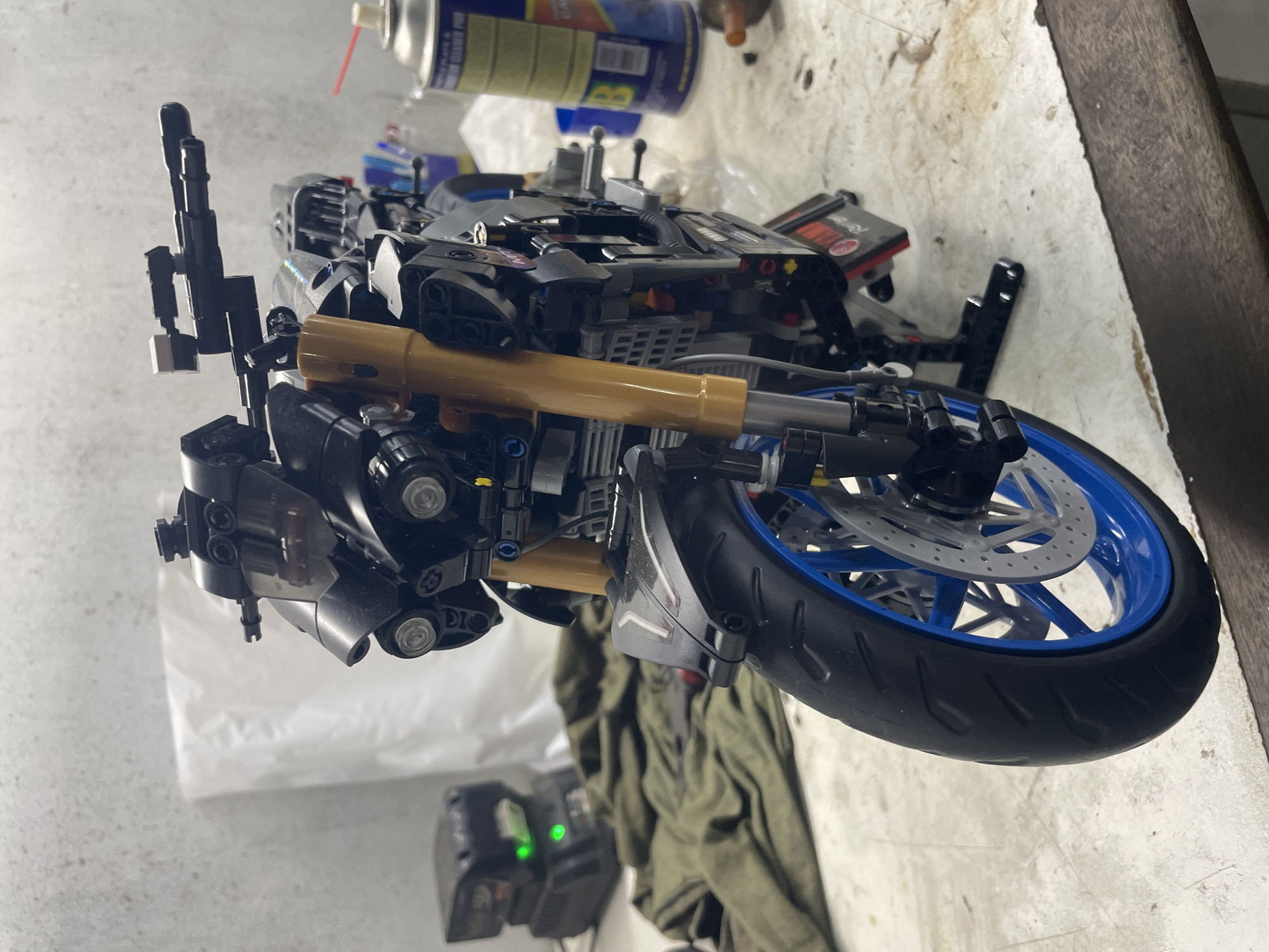 A detailed LEGO motorcycle model is the central subject of the image. It features black, blue, and gold-colored elements, depicting details like a front wheel, engine components, and handlebars. The motorcycle is positioned on a cluttered surface or workbench with tools and other objects in the background. Also on the work bench is a can of spray cleaner. A green garment partially covers the bottom of the frame. An electronic device with two illuminated green lights rests at the lower left. The environment suggests an indoor workshop or hobby space. There is no information regarding Bac Nha Trang Ward, Vietnam in this image.