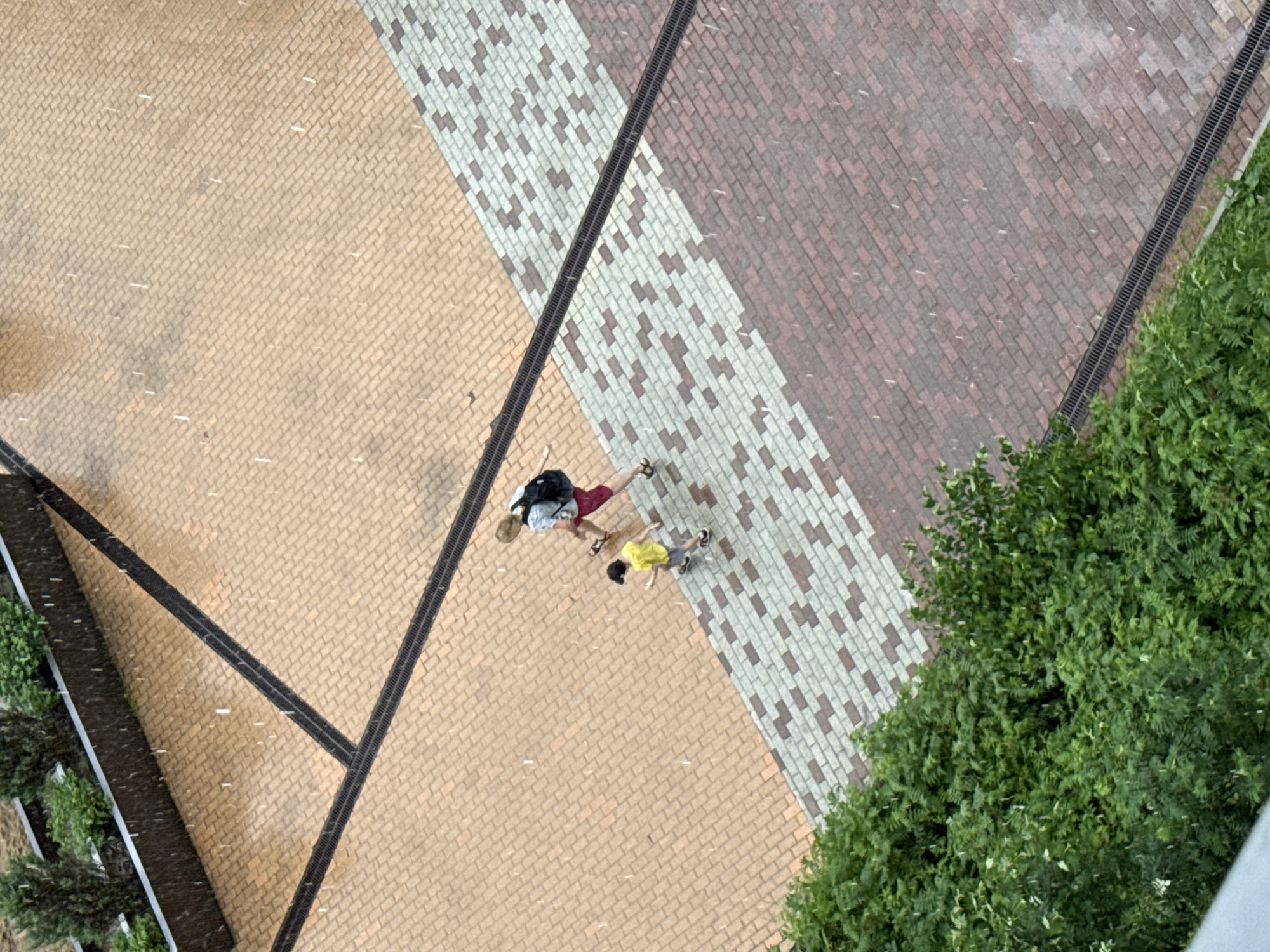 This is an aerial, medium shot taken on a rainy day. Two people, an adult and a child, are walking on a paved pathway. The adult, wearing a light-colored shirt and dark shorts, carries a backpack. The child, in a yellow shirt and dark shorts, walks slightly ahead. The pathway is laid with rectangular bricks in a pattern of tan, light gray, and dark red. A dark grate runs along the edge of the path. Lush green bushes are visible in the lower left corner. The scene is set in an outdoor urban environment, likely a park or residential area in Moscow, Russia.