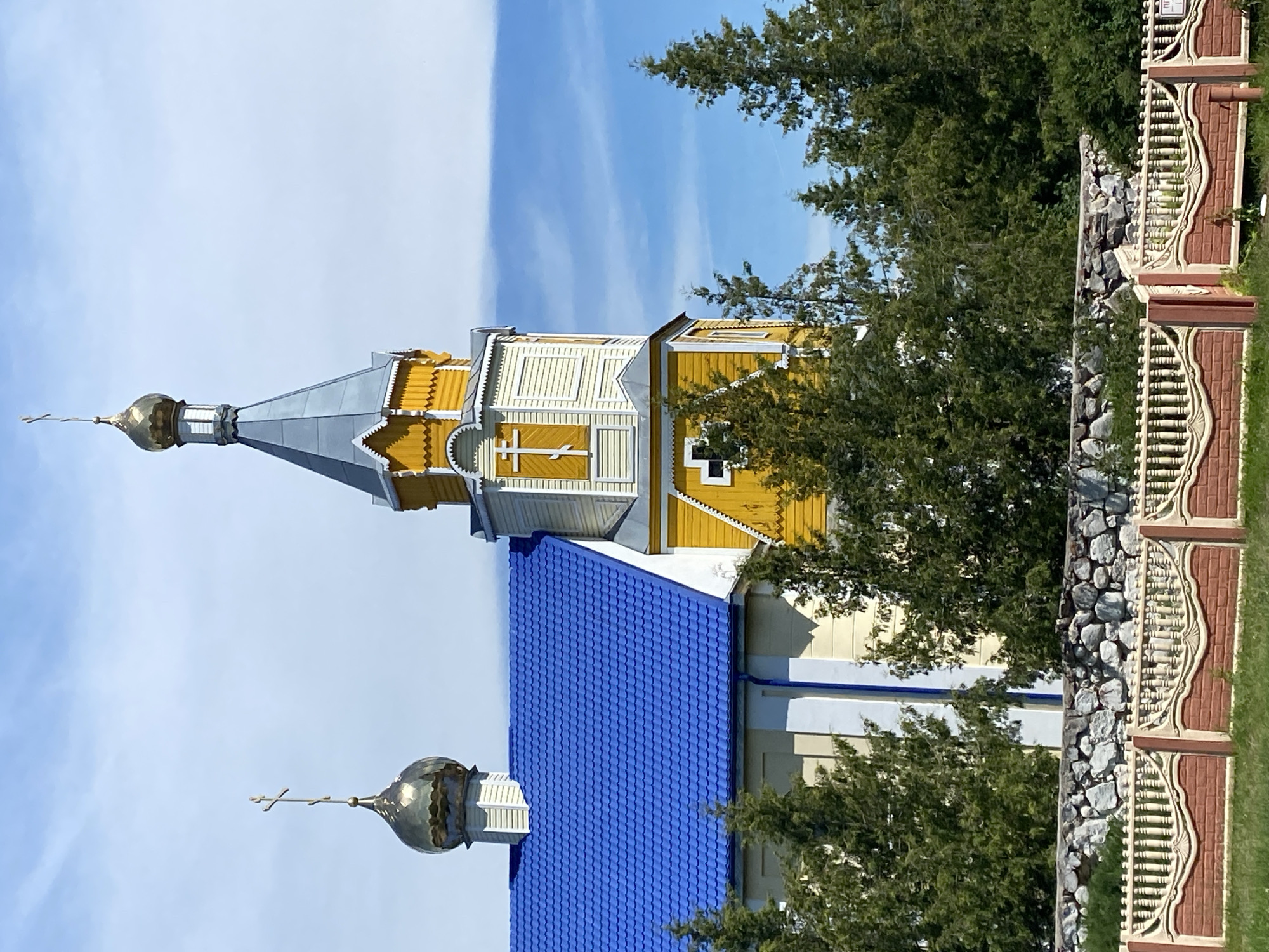 The image captures a yellow wooden church with a vibrant blue roof and a golden dome topped with a cross. A smaller bell tower, also yellow and adorned with crosses, stands adjacent to the main structure. The church is partially obscured by lush green trees. In the foreground, a stone wall separates the churchyard from a decorative fence with brick and concrete elements. The sky above is a clear blue with scattered white clouds. The scene is set in the village of Volchinski in the Volchinski Selsoviet, Belarus.