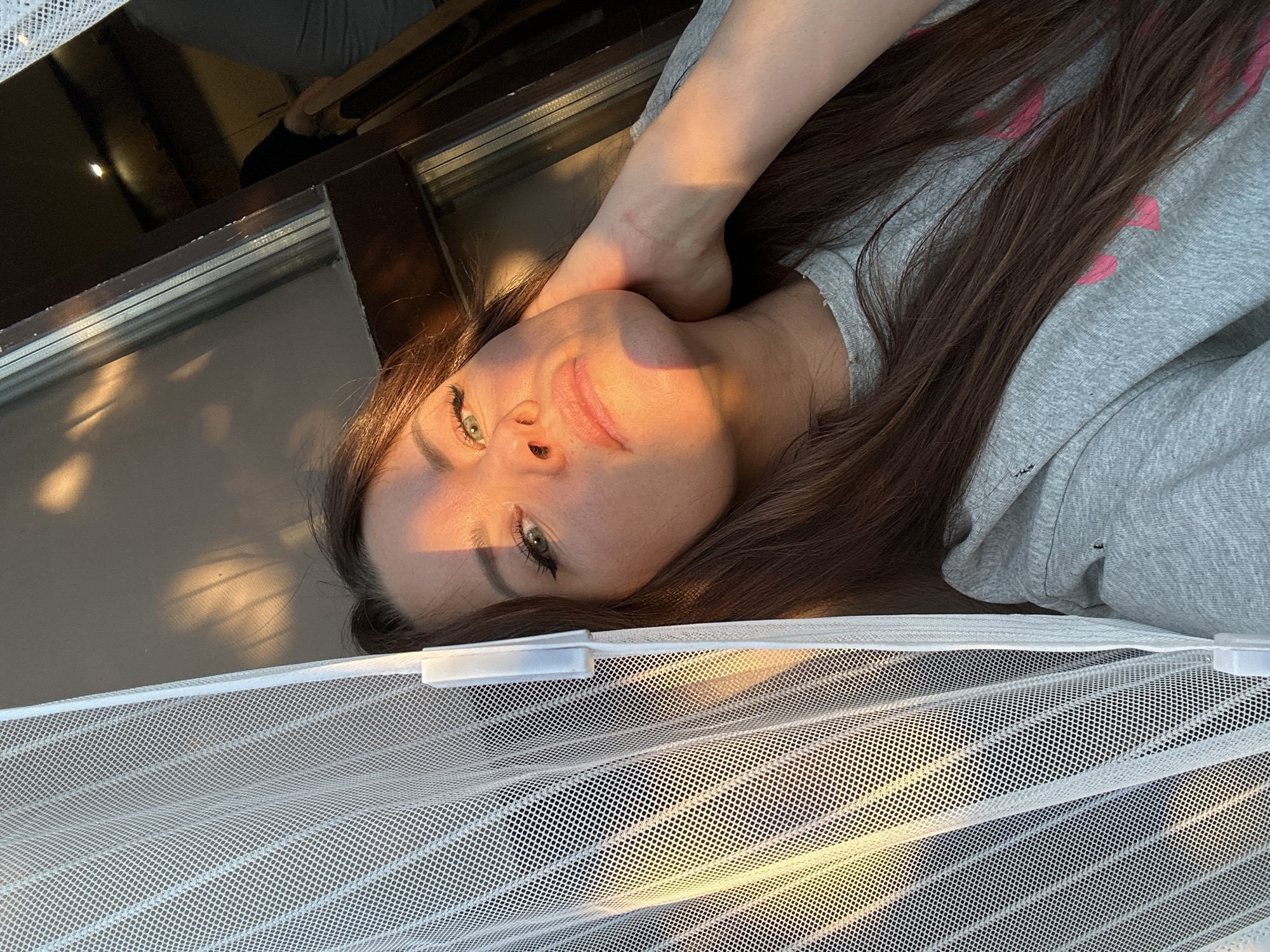 The image features a woman with long, dark brown hair and green eyes, illuminated by warm, golden sunlight. She is lying down, looking towards the camera with a calm and gentle smile, her right hand resting near her chin. Her fair skin is bathed in the soft glow, suggesting the time of day is either sunrise or sunset on a sunny day.
She is wearing a light grey long-sleeved top adorned with faint pink patterns that resemble hearts. The setting appears to be indoors, possibly near a window or doorway. A dark brown window frame is visible behind her head, reflecting some ambient light. In the foreground, a white mesh screen with a fine, repetitive grid pattern partially obstructs the view, with light filtering through it and casting subtle shadows. This screen likely serves as a mosquito net or a privacy curtain. The overall impression is one of peaceful serenity. The location context provided is Chaikovsky, Russia.
