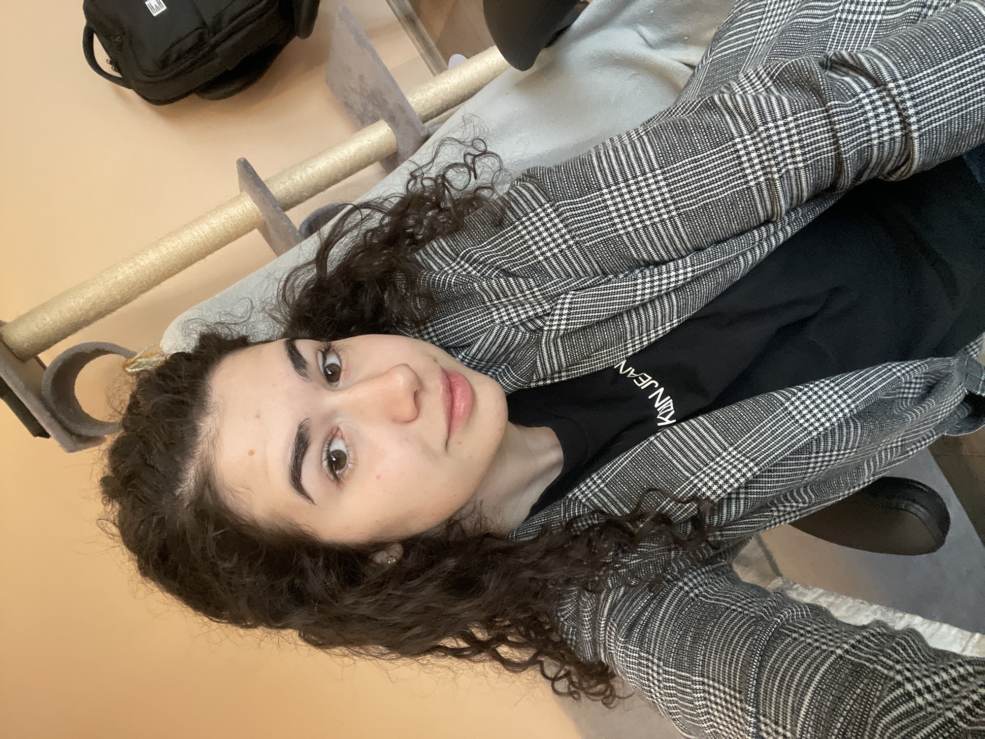 A person with curly dark hair and a fair complexion is lying on a surface, likely a bed or a couch, wearing a black T-shirt with white lettering and a grey and white plaid blazer. The T-shirt displays the partial text "KLIN JEAN". A black backpack is visible in the upper left corner, resting on a light-colored surface. Parts of a cat tree or scratching post are visible near the person. The background wall is a light peach color. The location in the city of Sofia, Bulgaria is not discernible from the image.