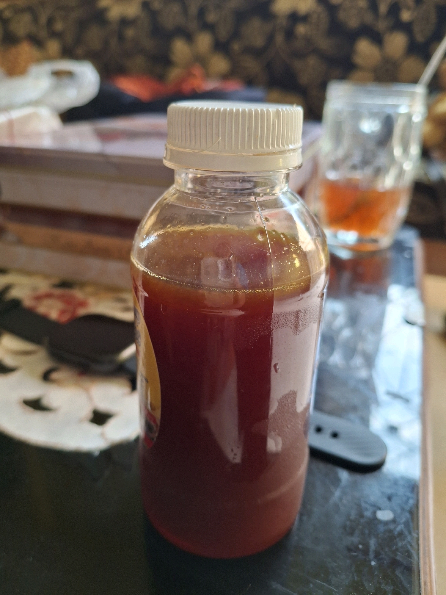 A glass bottle with a white cap contains a dark red liquid. The bottle is sitting on a dark, reflective surface, possibly a table. In the background, out of focus, there is a stack of books or boxes. To the right of the bottle, there is a glass with a lighter red liquid and a straw. A black watch strap is visible on the surface in front of the bottle. The lighting suggests an indoor setting. No people are visible. This scene is not specifically indicative of Lumajang, Indonesia; it's a generic indoor setting.