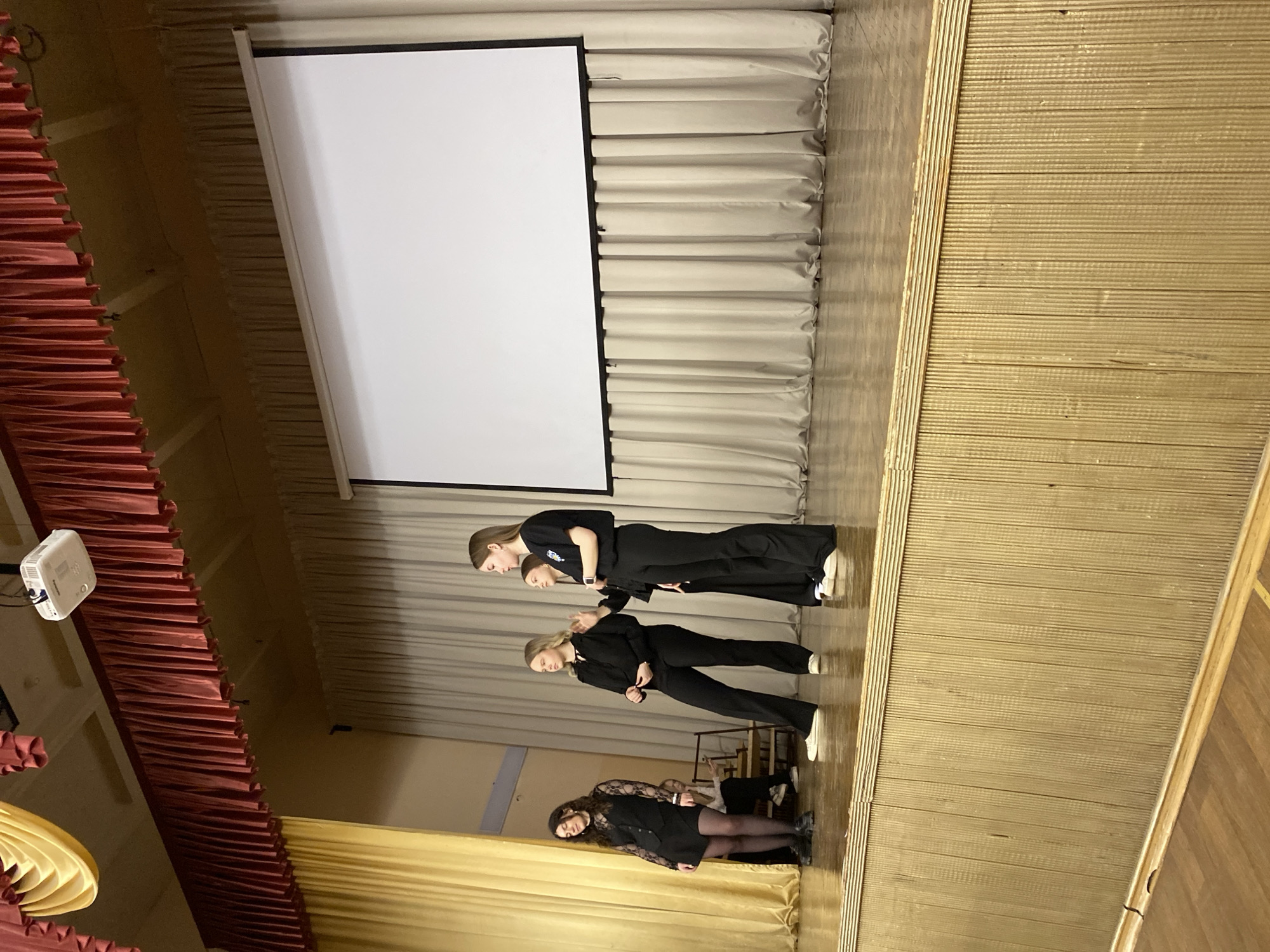 Three young women stand on a stage in Votkinsk, Russia, beneath a projector screen. The women are dressed in dark clothing, with the woman closest to the foreground wearing a dress with sheer lace sleeves, and the other two dressed in black pants and tops. The stage is decorated with maroon and yellow drapery along the sides. The stage floor and the walls behind the drapery are light-colored. A projector is mounted on the left.