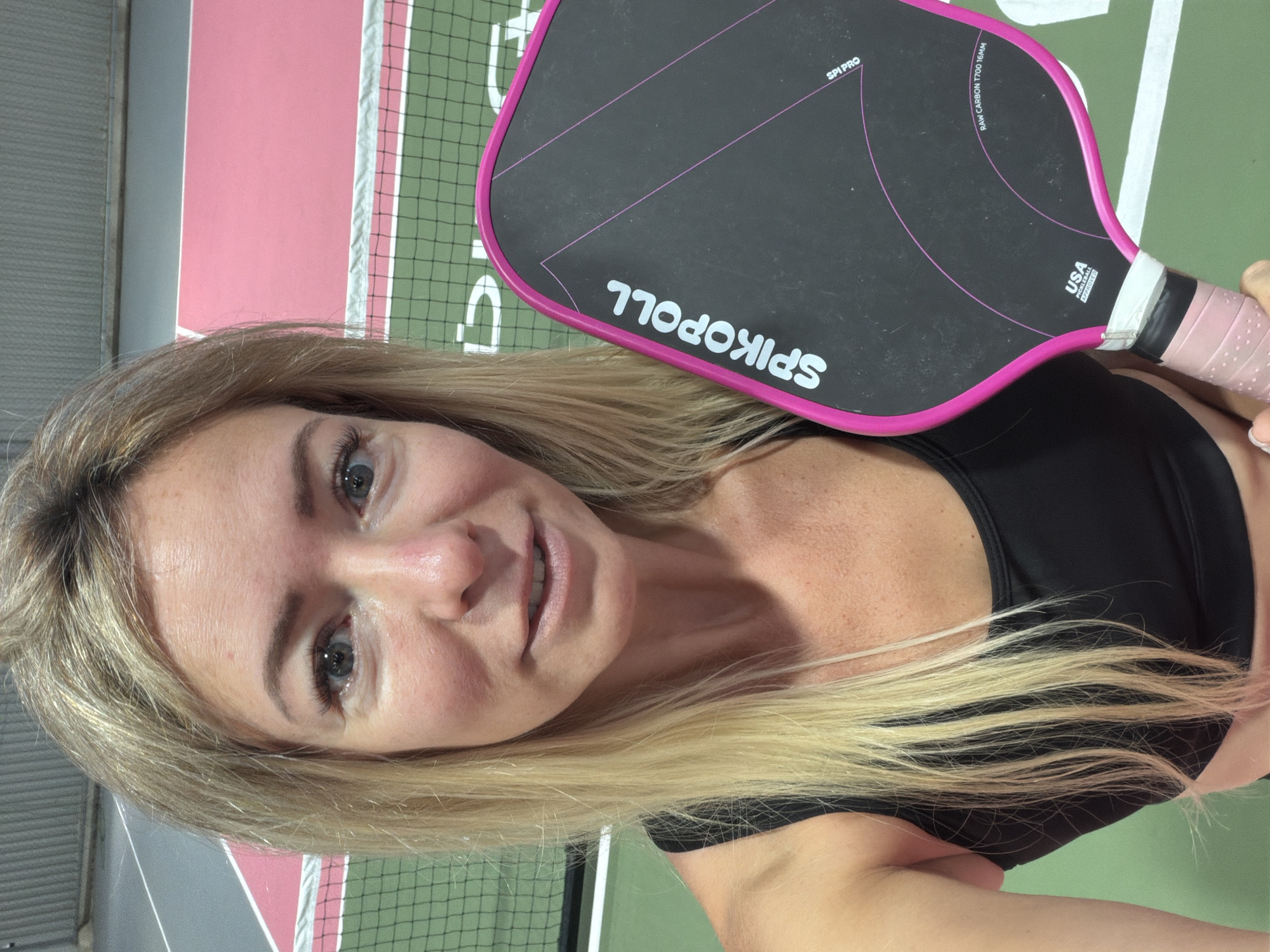 A blonde woman is taking a selfie indoors on a pickleball court. She is holding a black and pink pickleball paddle with "SPIKOPOLL" written on it. She is wearing a black sports bra. The court has green and pink surfaces and a net. There is no information in the image to determine its location in Da Lat, Vietnam.