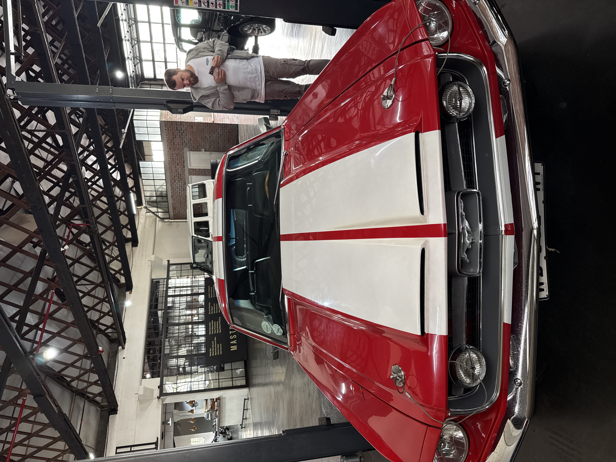 An image, rotated counter-clockwise, captures an indoor automotive setting, possibly a workshop or showroom, with an industrial architectural style. The primary subject is a classic red Ford Mustang car, featuring distinctive white racing stripes running down its hood. Its front bumper, grille, and headlights are prominently displayed, with the Mustang emblem visible on the grille.
Standing next to the Mustang, on the same car lift platform, is a man with a beard, dressed in a grey hooded top, a white t-shirt, and light brown trousers. He is engrossed in his smartphone. Above him, on an upper section of the car hoist, a dark-colored vehicle, possibly a larger car or SUV, is partially visible.
The background showcases the building's interior, featuring high ceilings with exposed dark metal trusses and large windows with grid patterns, allowing ample natural light, indicating it is daytime. A large black banner on a distant wall lists various automotive services, with phrases such as "Wet Collection," "Deep Clean," "Paint Protection," and "Mag Wheel Polishing" clearly legible, along with "SERVICES" at the bottom. The letters "MAST" are also visible on the banner. The setting suggests a specialized automotive facility, potentially in Cape Town, South Africa, dedicated to car detailing, maintenance, or display.