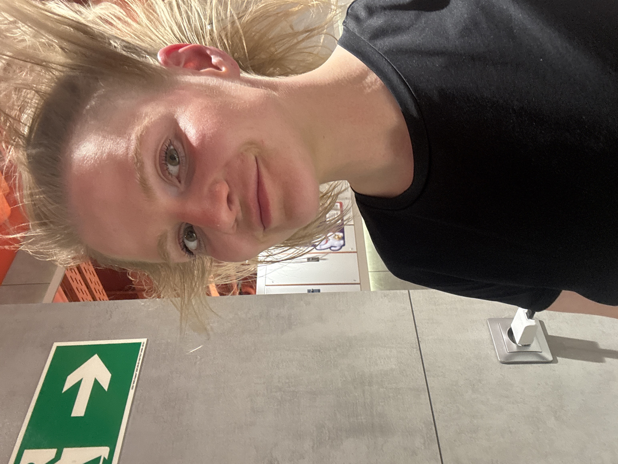 A woman with blonde hair and a black shirt is photographed indoors, possibly in a commercial or industrial space. An illuminated exit sign is visible, and there is an orange structure in the background. A white electrical outlet is visible in the foreground. The location in the city of Saint Petersburg, Russia, cannot be determined from the information presented in the image.