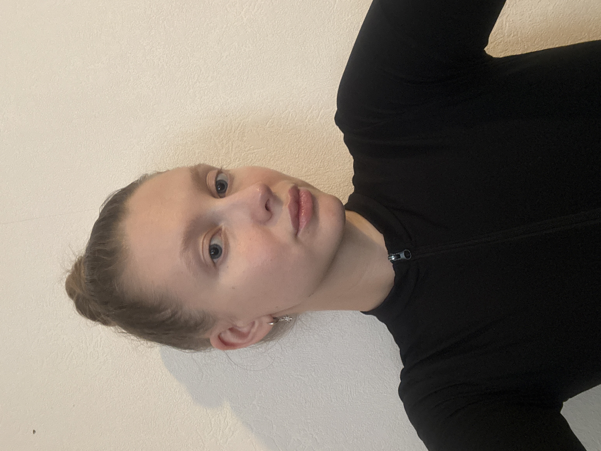 A young adult female, positioned for a selfie, is depicted indoors in the city of Izhevsk, Russia. She has fair skin, light blue eyes, and light-colored eyebrows, with her brown hair pulled back into a bun atop her head. She wears a plain black, long-sleeved top featuring a short zipper at the neck. A small silver hoop earring with a dangling charm is visible in her left ear. The background is a textured, off-white wall, illuminated by an unseen light source that casts a soft shadow to her left.