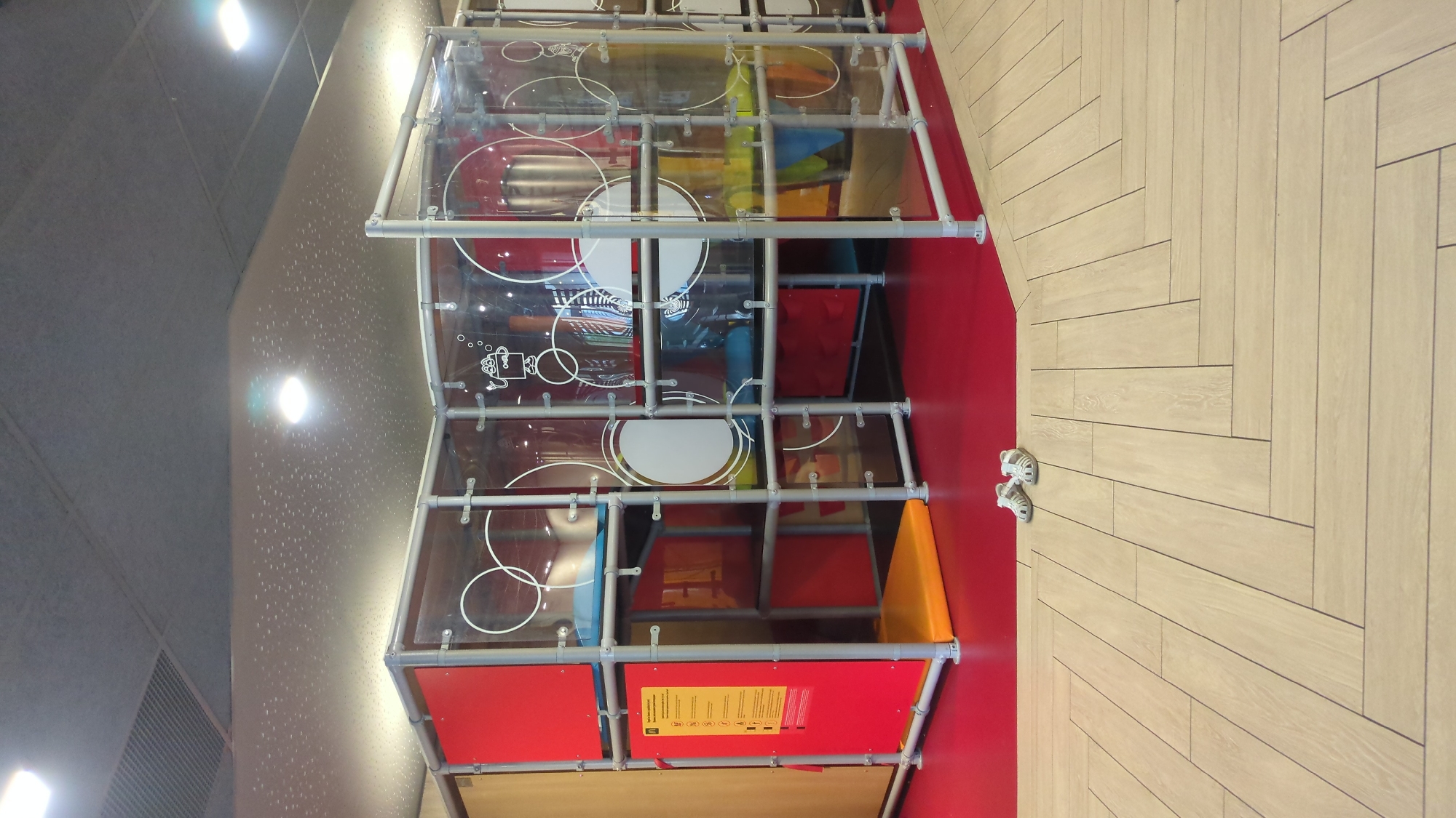 This image depicts an indoor children's play structure, likely located in a McDonald's restaurant, as suggested by the prominent red panel with the McDonald's logo and informational signs. The structure is made of clear plastic panels supported by a metal frame. Inside the play area, there are multiple levels with brightly colored surfaces, including red, blue, yellow, and orange mats. The floor of the main area is red, while the foreground shows a floor with light-colored wooden planks laid in a herringbone pattern. A pair of white children's sandals are visible on the wooden floor near the edge of the red play area. The ceiling is gray and appears to be made of acoustic tiles, with several circular light fixtures. The overall scene is clean and well-lit, indicating a public indoor space designed for children's recreation. The specific location in Chișinău, Moldova, cannot be determined from the visual information provided in the image.