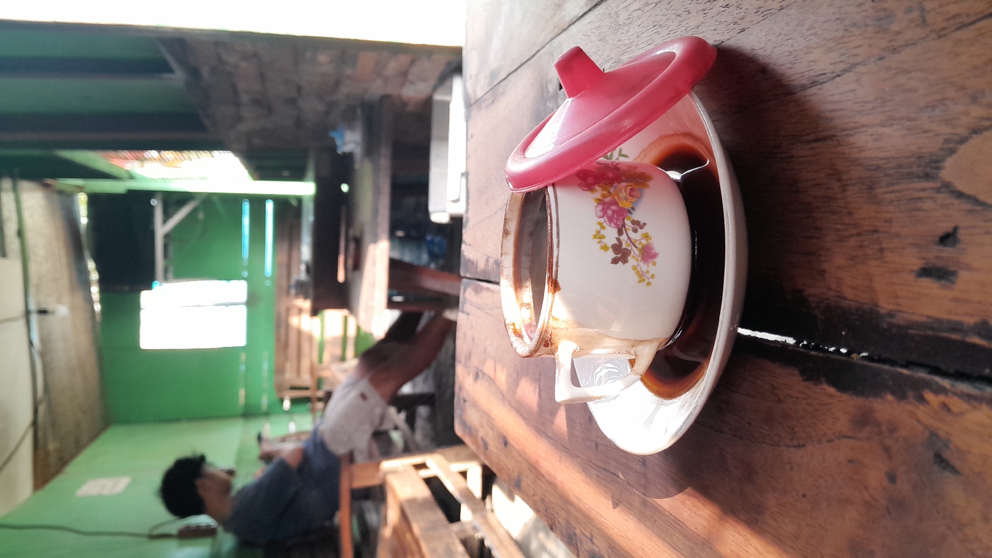 In Balun, Indonesia, a close-up shot focuses on a white ceramic cup filled with black coffee, resting on a matching saucer. The cup is adorned with a floral pattern featuring pink and yellow flowers. A bright pink lid is placed beside the cup. Both the cup, saucer, and lid are positioned on a wooden table with visible grain and imperfections. In the background, a person wearing a blue shirt and white shorts is seated on a wooden bench. The environment appears to be an outdoor or semi-outdoor area with green walls and a corrugated roof. Sunlight streams through a window, casting shadows across the table. The scene suggests a casual, possibly rustic, setting.