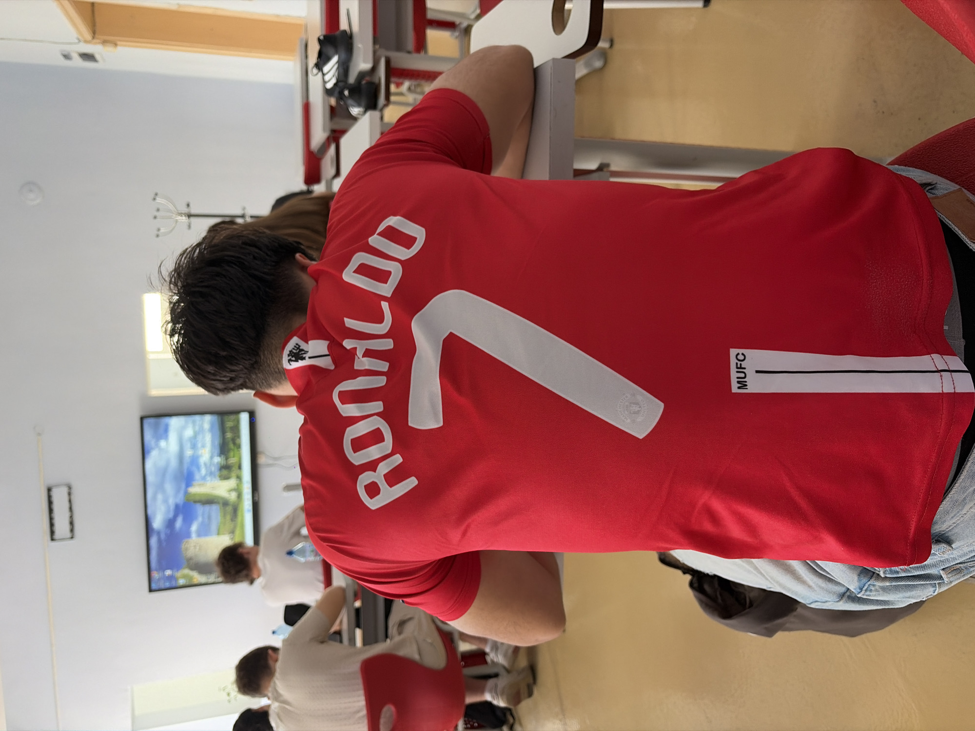 The image depicts a classroom setting with several students seated at desks. The main subject is a student in the foreground, facing away from the camera, wearing a red t-shirt with "RONALDO" and the number "7" printed on the back. A logo, likely a football club crest, is visible on the t-shirt. Other students are visible in the background, also seated at desks, some looking towards a large screen displaying a landscape image of a castle. Desks, chairs, and a whiteboard are present in the room. The flooring is a light yellow. The environment appears to be a school or educational institution. No specific details about the city of Plovdiv, Bulgaria are present in the image.