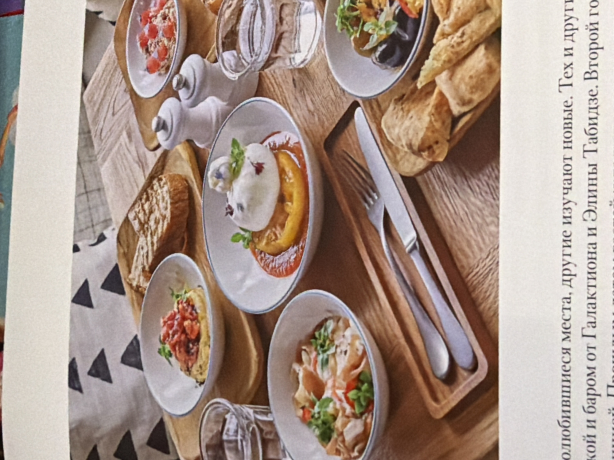 The image shows a photograph of a spread of food on a wooden surface, with plates of various dishes, including what appears to be bread, salad, and a tomato and cheese dish. Cutlery is also present. The image is taken at an angle, and a portion of the frame includes text in Russian.