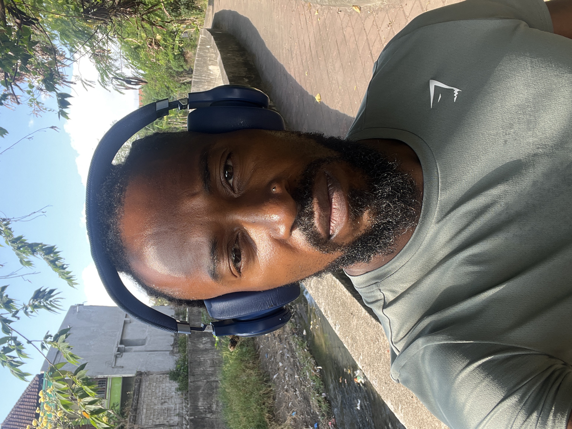 In Denpasar, Indonesia, a Black man with dark skin, short hair, and a full beard is pictured outdoors, wearing large navy blue headphones and a dark olive green t-shirt with a white logo on the left chest. He is looking directly at the camera. He is positioned near a concrete walkway, with a narrow, somewhat polluted canal to his left. The canal has some debris in it, and the bank is overgrown with grass. Behind the canal, a grey concrete wall and a multi-story building with a green lower section are visible. To the right of the man, the concrete walkway continues, bordered by a low, weathered concrete wall. Trees and foliage with sunlight filtering through them are present in the background, creating shadows on the walkway. The sky is blue with some white clouds. The overall impression is a casual outdoor setting in an urban environment.