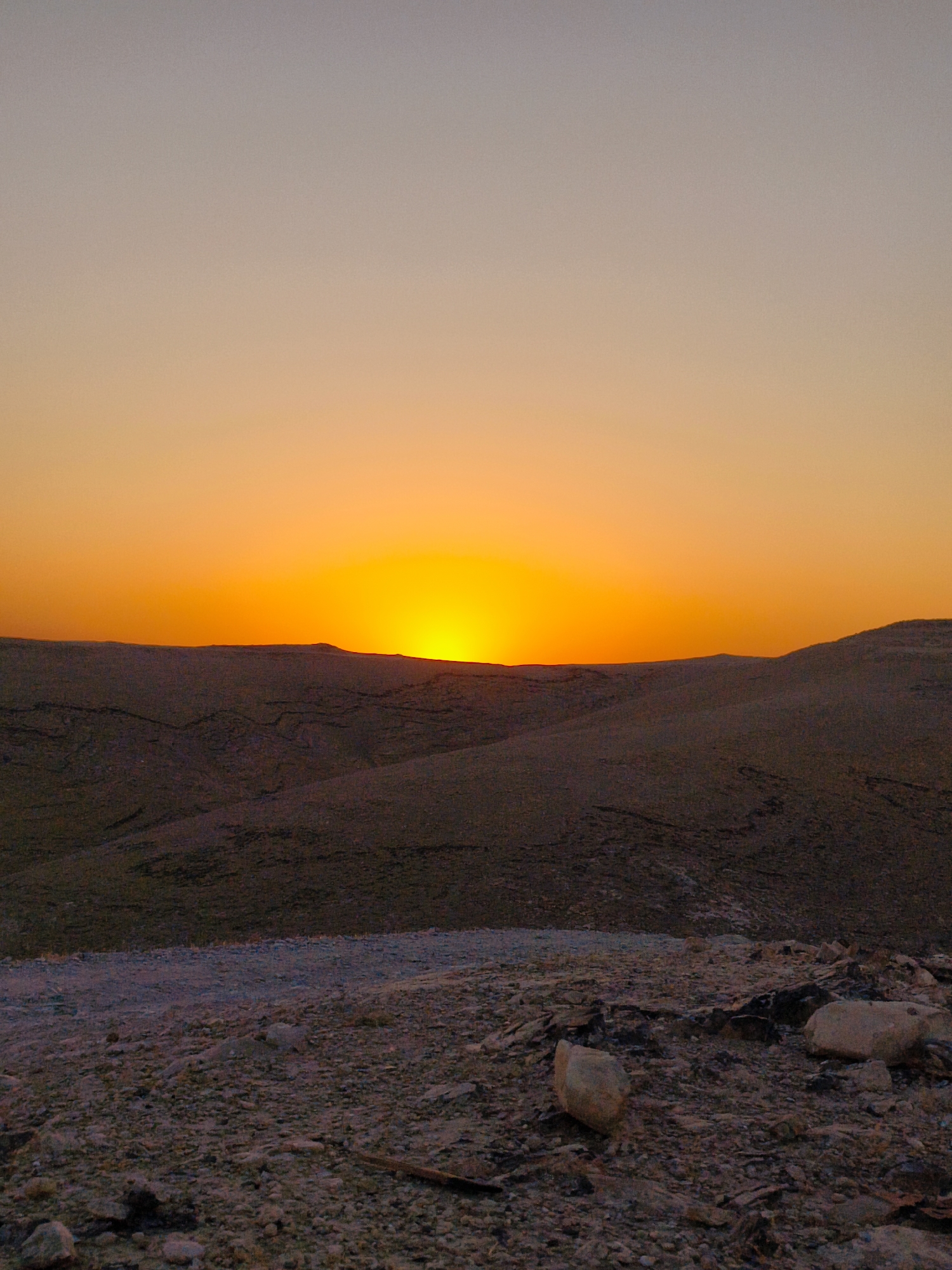 The image depicts a sunset over a rocky, arid landscape in Keidar, Palestinian Territories. The sky transitions from a pale grey at the top to a vibrant orange near the horizon, where the sun is setting. Below the sky, rolling hills are visible, appearing dark and textured. The foreground is covered in small stones and gravel, with several larger rocks scattered across the ground. There are no people or structures visible in the image. The overall impression is one of natural, undisturbed wilderness.