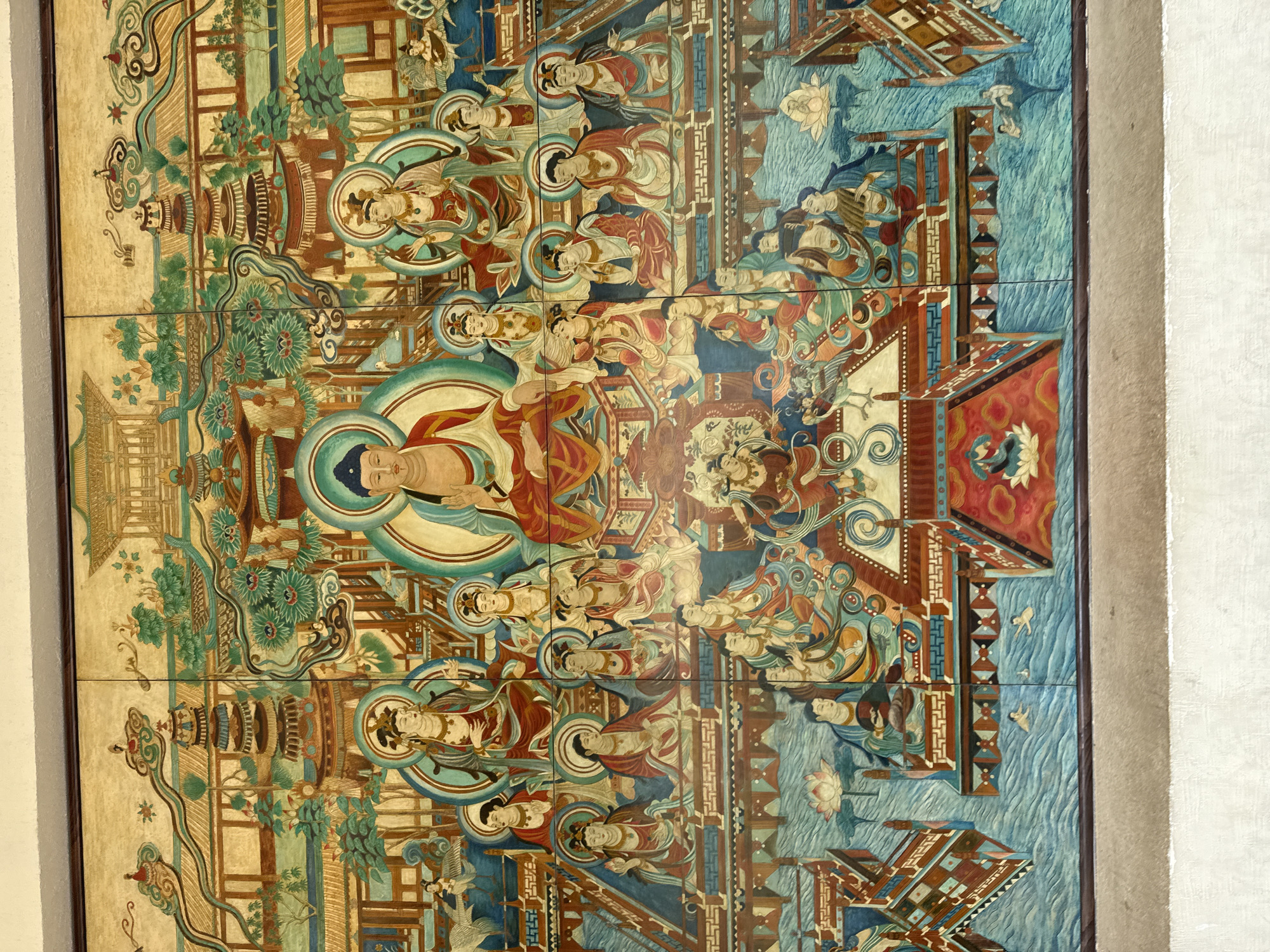 The image is a section of wall art comprised of square panels, likely tiles, depicting a scene of Buddhist figures and traditional Chinese architecture.
**People:** Numerous figures with serene expressions and halo-like circles behind their heads. They are dressed in traditional robes and are positioned in poses suggesting reverence and meditation.
**Subjects:** The central figure is larger and more prominent, likely representing Buddha. Smaller figures surrounding him may represent Bodhisattvas, disciples, or other celestial beings. The scene also includes architectural elements such as pagodas, pavilions, and decorative bridges. There are also depictions of water, lotus flowers, and other symbolic flora and fauna.
**Objects:** The objects include robes, halos, buildings, water, plants and lotus flowers
**Actions:** The figures are engaged in activities of worship, meditation, or teaching.
**Interactions:** The figures are interacting with each other through gestures, expressions, and their relative positions in the scene. The architectural elements and natural features contribute to the overall sense of harmony and spiritual significance.
**Environment:** Based on the style of art and architecture, this image most likely depicts a scene from the Buddhist caves in Dunhuang City, China.