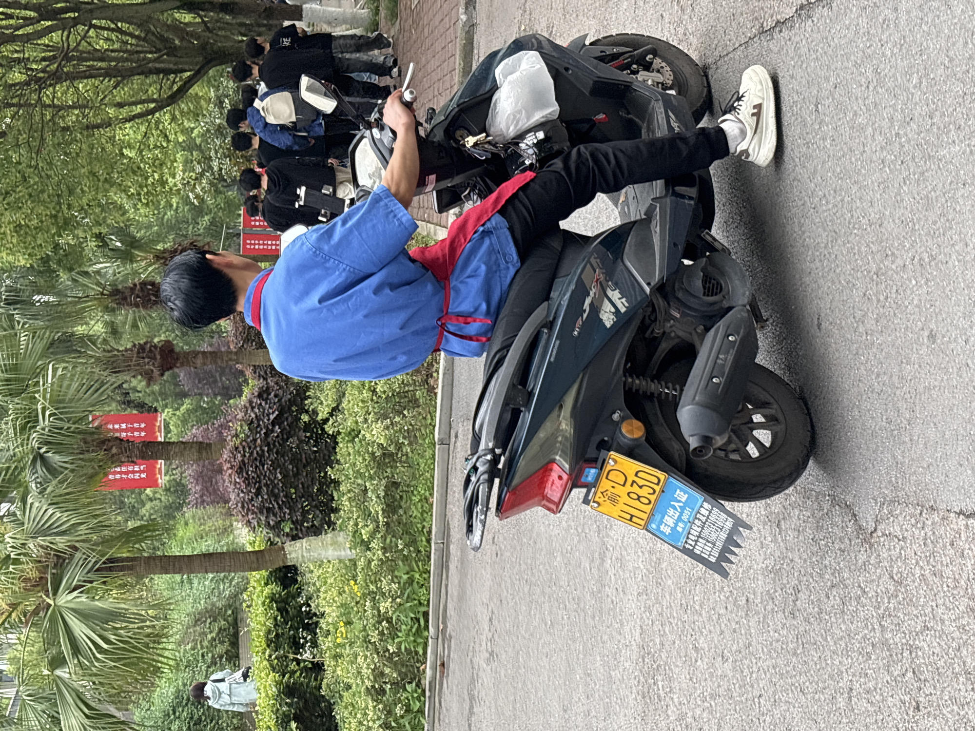 A man on a dark-colored scooter, license plate "渝・D H183D", is parked on a paved surface next to greenery in Jiangjin, China. He is wearing a blue short-sleeved shirt with red trim and black pants, and white sneakers. In the background, other people stand along the edge of the road. Signboards with Chinese characters are visible amongst palm trees and shrubbery in the vicinity.
