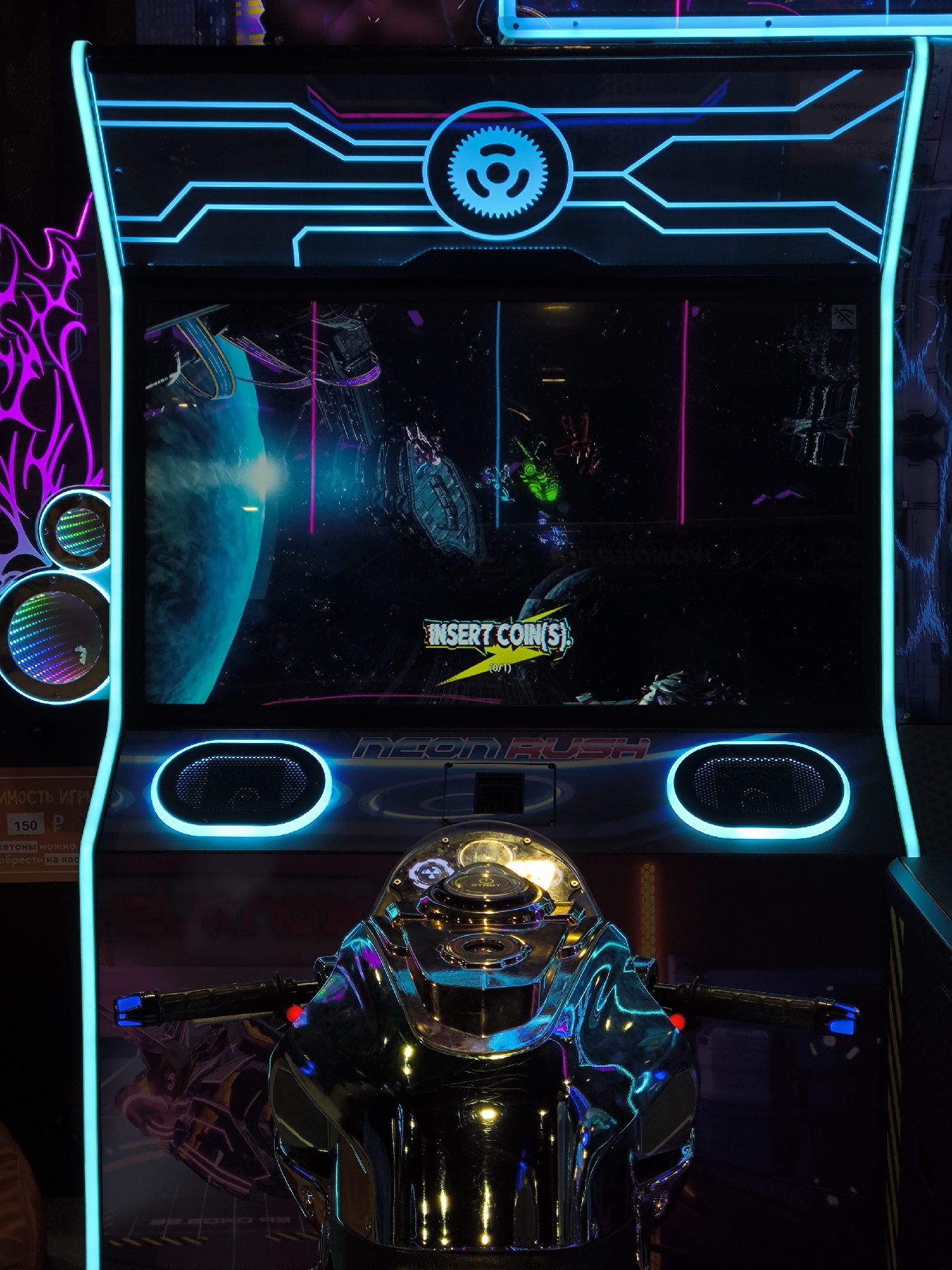 An arcade game cabinet for "Neon Rush" is shown in this vertical, close-up shot. The cabinet features a large screen displaying a futuristic racing game with neon blue and pink accents. The front of the cabinet has a chrome-finished motorcycle steering column and handlebars. Two circular speakers with blue lighting are located on either side of the screen, and the entire cabinet is outlined with neon blue lights. The background is dark and appears to be an indoor setting, possibly an arcade or entertainment center. There are no people visible in the image. The lower left corner of the cabinet has some text and numbers printed on it, including "150 Р".