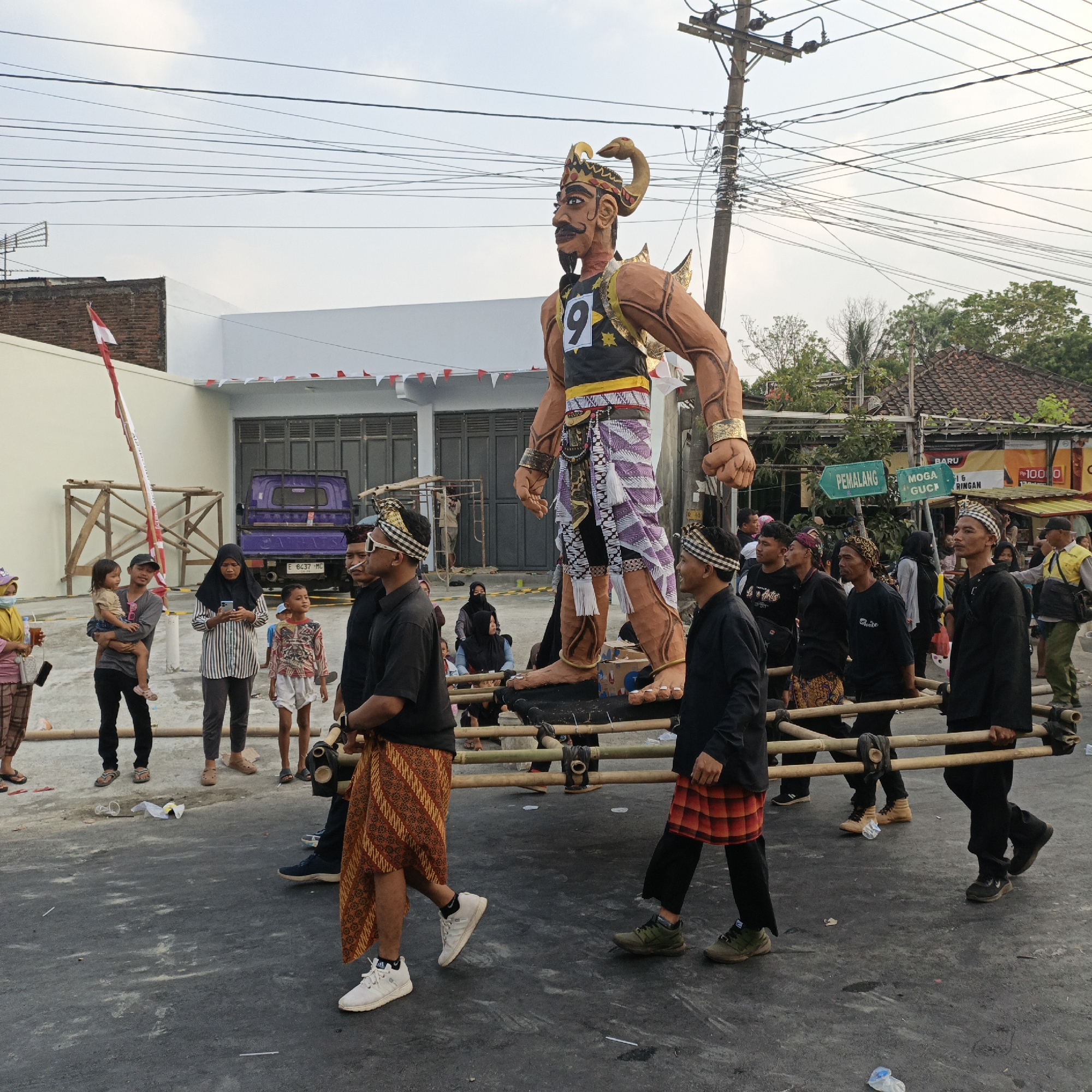 In Randudongkal, Indonesia, a large, anthropomorphic effigy, possibly a representation of a mythical giant or warrior, is being carried on a bamboo platform by a group of men. The effigy is adorned with a headdress featuring horns and a snake-like element, and wears a black vest with the number "9" prominently displayed, along with a patterned sarong. The men carrying the effigy are dressed in black shirts and sarongs, some with traditional headbands. A purple utility vehicle is parked in the background, along with buildings and electrical poles with numerous wires crisscrossing the sky. Several spectators, including families with children, are observing the procession. Red and white triangular flags are strung across the scene, suggesting a festive or nationalistic event. The ground is paved and shows some debris, including scattered plastic bottles. The sky is overcast, indicating a possibly late afternoon or early morning setting.