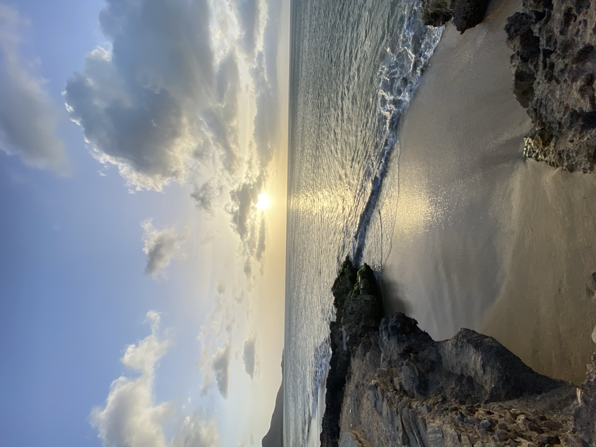 The image captures a sunset view at a beach in Juan Griego, Venezuela. The sky is filled with clouds, partially obscuring the sun. The water reflects the sunlight, creating a shimmering effect. Rocks are visible in the foreground on the right and in the middle of the image, and a mountain appears in the distance on the left. The beach is sandy, and gentle waves are lapping at the shore. The image is rotated 90 degrees counter-clockwise.