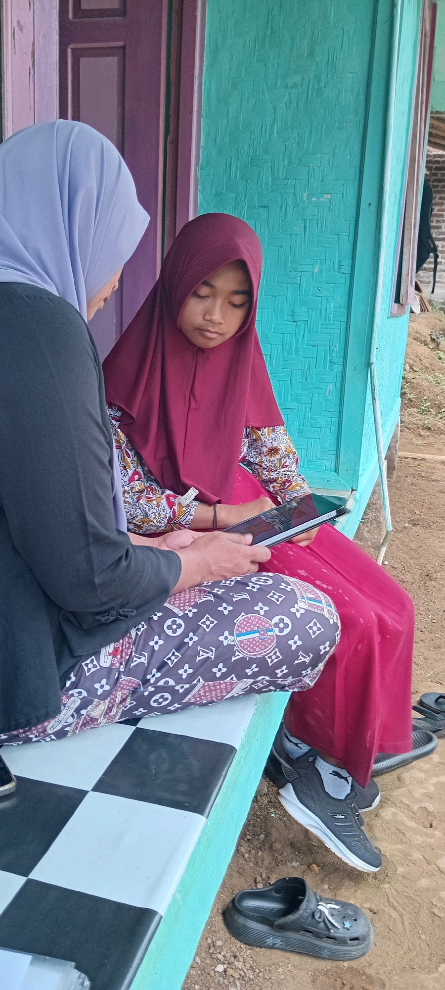 In a rural outdoor setting, possibly in the city of Sodonghilir, Indonesia, two young females are seated on a checkerboard-patterned bench. The female on the right wears a maroon hijab, a patterned shirt with red and white floral motifs, and a red skirt. She is looking down at a tablet device held in her hands. The female on the left wears a light blue hijab and a dark grey top. Her patterned pants feature a repeating Louis Vuitton-inspired motif of interlocking "L" shapes and quatrefoils in maroon and white on a dark background. Her hands are also near the tablet. The background consists of a turquoise-painted wooden wall with horizontal planking, and a purple-painted wooden door frame. The ground is composed of loose dirt and small stones, with a pair of black clogs and a black sandal visible on the ground to the right.