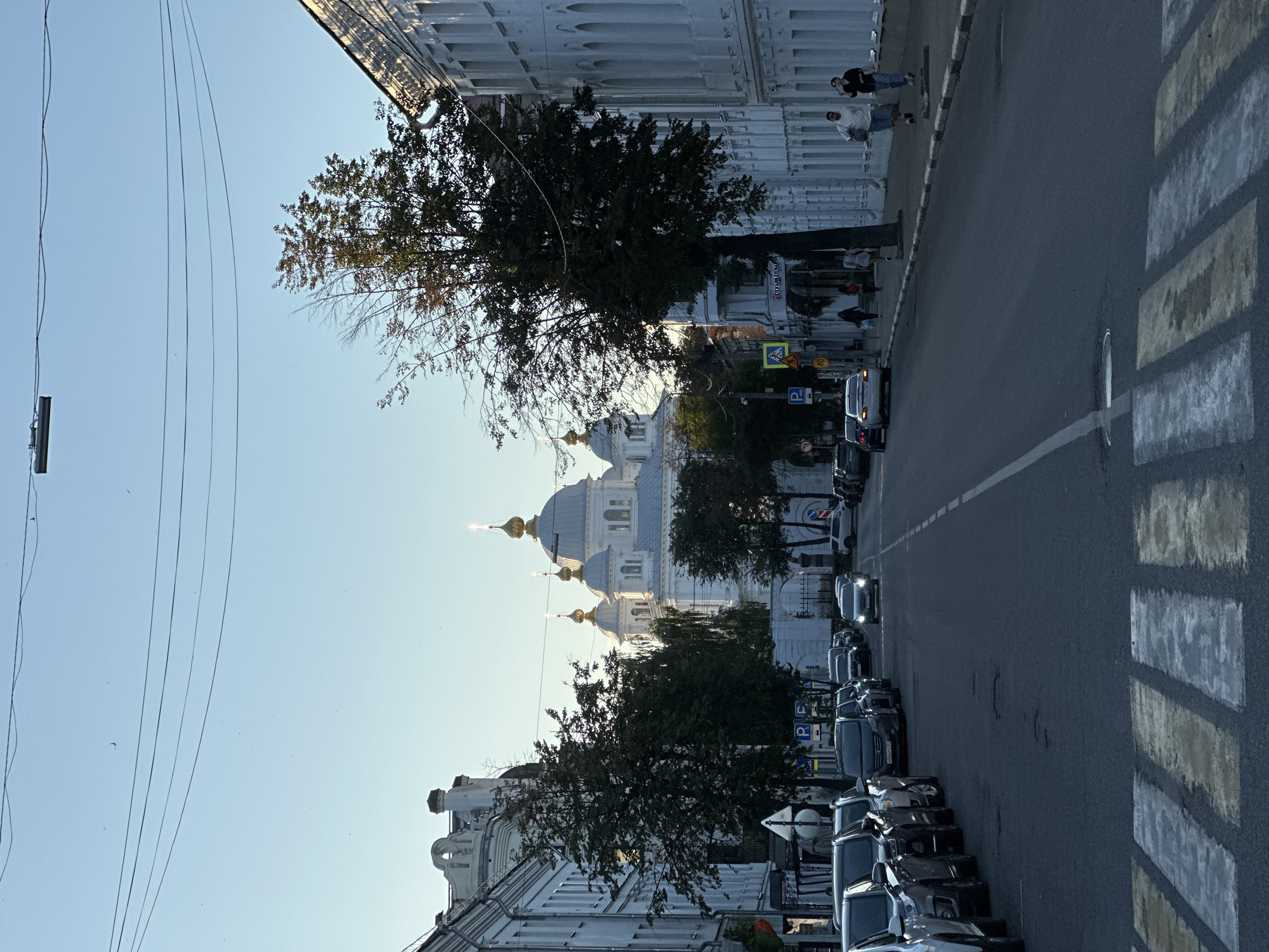The image shows a street scene in Yazvitsevo, Russia, with a white domed church visible in the background. The church has several golden domes and spires, and is surrounded by trees. On the right side of the street, there are white buildings with arched windows. On the left side of the street, there are parked cars. The street has a pedestrian crossing in the foreground. Several people are walking on the sidewalk on the right side of the street. The sky is clear and blue, and the sun is shining. The scene appears to be set during the daytime, possibly in the late afternoon or early evening due to the lighting.