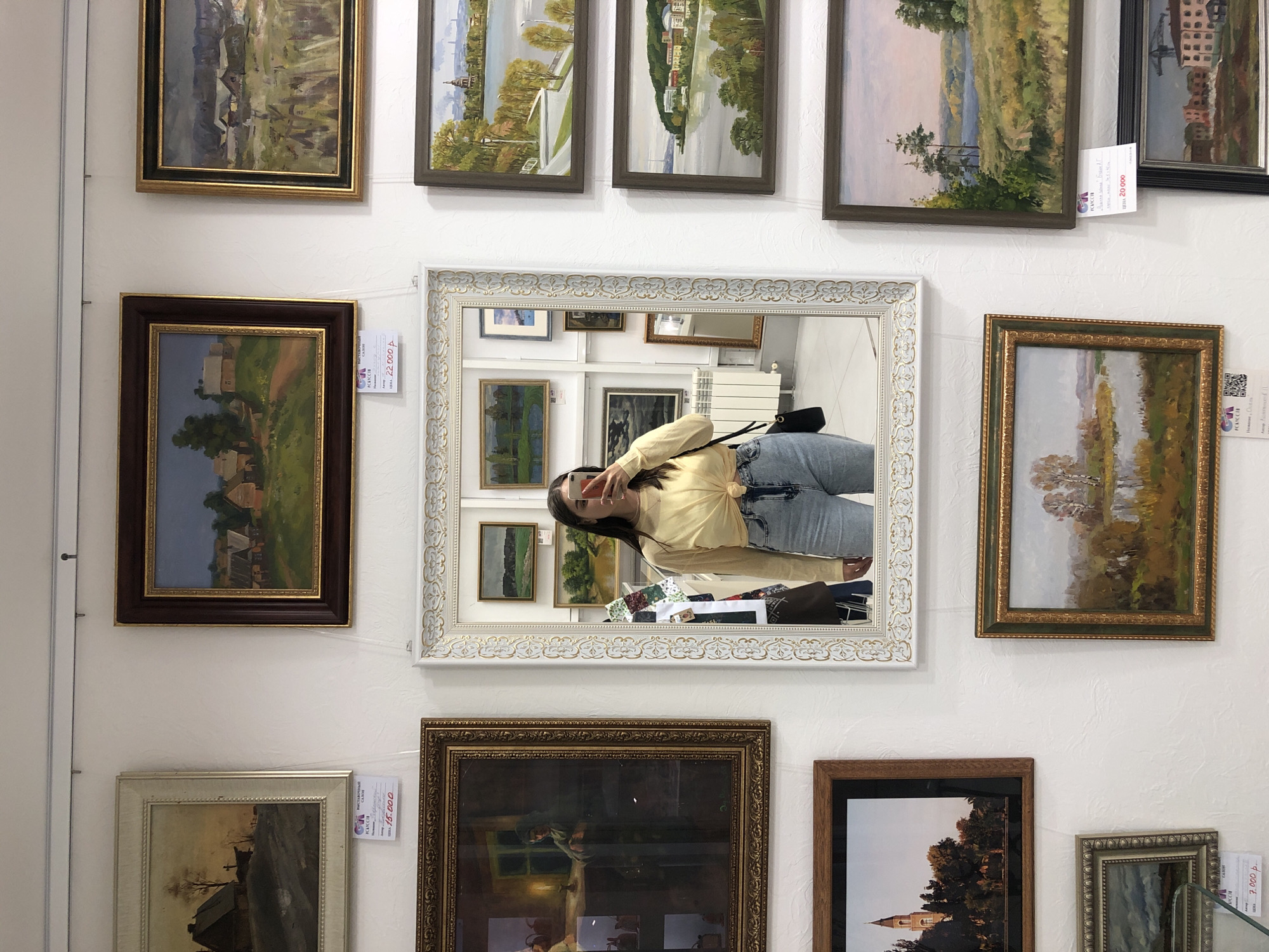 The image shows a wall in an art gallery or exhibition space, displaying a collection of framed paintings. The main subject is the display of artwork, with a woman taking a selfie in a large ornate mirror that is part of the display. She is wearing a yellow blouse and blue jeans and is holding up her phone. The paintings are varied in subject matter, including landscapes and village scenes. Several of the paintings have price tags attached, with visible text including prices in Rubles (e.g., "22 000 p.", "15 000", "7 000 p."). One tag also includes the word "ПЛАНСИР". The setting appears to be indoors, with white textured walls and wires visible for hanging the artwork. The lighting suggests it is daytime. The overall impression is one of an art exhibition, possibly in Izhevsk, Russia, given the mention of Rubles and the style of some of the paintings which might be indicative of Russian art.