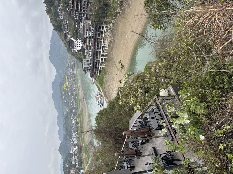 The image displays an elevated panoramic view of a river valley in Furong, China, under an overcast grey sky. In the immediate foreground, an empty outdoor terrace with multiple dark-colored tables and chairs, along with closed brown patio umbrellas, is visible. This terrace is partially framed by dense, varied green and brown foliage. The midground is dominated by a wide, light blue-green river, which shows extensive areas of exposed light brown riverbed on both banks, indicative of low water levels. Several small boats are docked together on the river's left bank.

On the steep right bank, a town comprised of numerous traditional-style multi-story buildings with white walls and dark roofs is built into the hillside. One prominent structure on the right bank features an open, arched lower level that extends over the exposed riverbed. Another cluster of similar buildings is visible on the opposing left bank. Distant hillsides on both sides exhibit patches of green and barren agricultural land, interspersed with fields displaying bright yellow blooms, consistent with rapeseed. A communication tower stands atop a distant hill on the left side of the scene. No individuals are visible in the image.