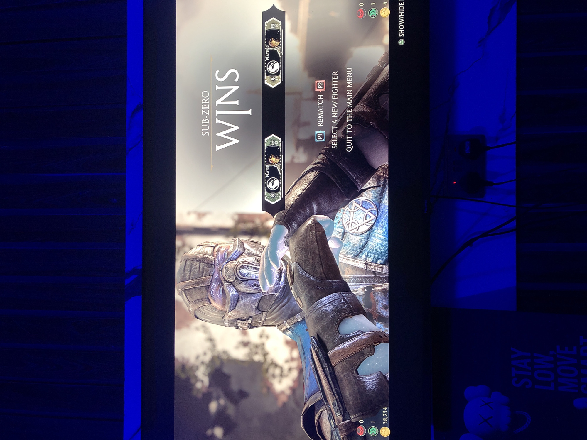 An image, rotated 90 degrees counter-clockwise, displays a video game screen showing the post-match victory screen for the character Sub-Zero from Mortal Kombat. The screen prominently features "SUB-ZERO WINS" in large, stylized text. Below this, user interface elements indicate player one (P1) has achieved one win, while player two (P2) has zero wins. Options are presented to the players: "P1 REMATCH P2," "SELECT A NEW FIGHTER," and "QUIT TO THE MAIN MENU." A "SHOW/HIDE" option with an 'A' button prompt is also visible at the top right, alongside numerical values like "0", "3", and "4." At the bottom of the screen, a score of "38,254" is displayed.
The game character, Sub-Zero, is depicted in detail, wearing intricate blue and silver armor with glowing ice-like hands. The screen is set within a darkened room, illuminated primarily by the monitor's glow and distinct blue LED strip lighting running vertically along the edges of the viewing area. On the right side of the image, an electrical outlet with plugged-in cables and a red indicator light is visible against the dark blue background. Further down on the right, a dark object or surface displays white text that reads "STAY LOW. MOVE SMART." next to a stylized character with 'XX' for eyes, resembling a KAWS Companion figure. The overall scene suggests a gaming session taking place in a dimly lit, possibly personal, space in Akwanga, Nigeria.