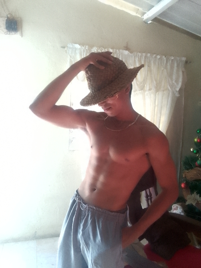 A shirtless man poses indoors with a woven hat partially obscuring his face. He has a slender build with visible abdominal muscles and wears light blue sweatpants. A silver chain is around his neck. One hand rests on his head, securing the hat, while the other is in his pocket. Behind him are white curtains and a portion of a decorated Christmas tree. The wall is a faded off-white, and an electrical fixture is visible in the upper left corner.
