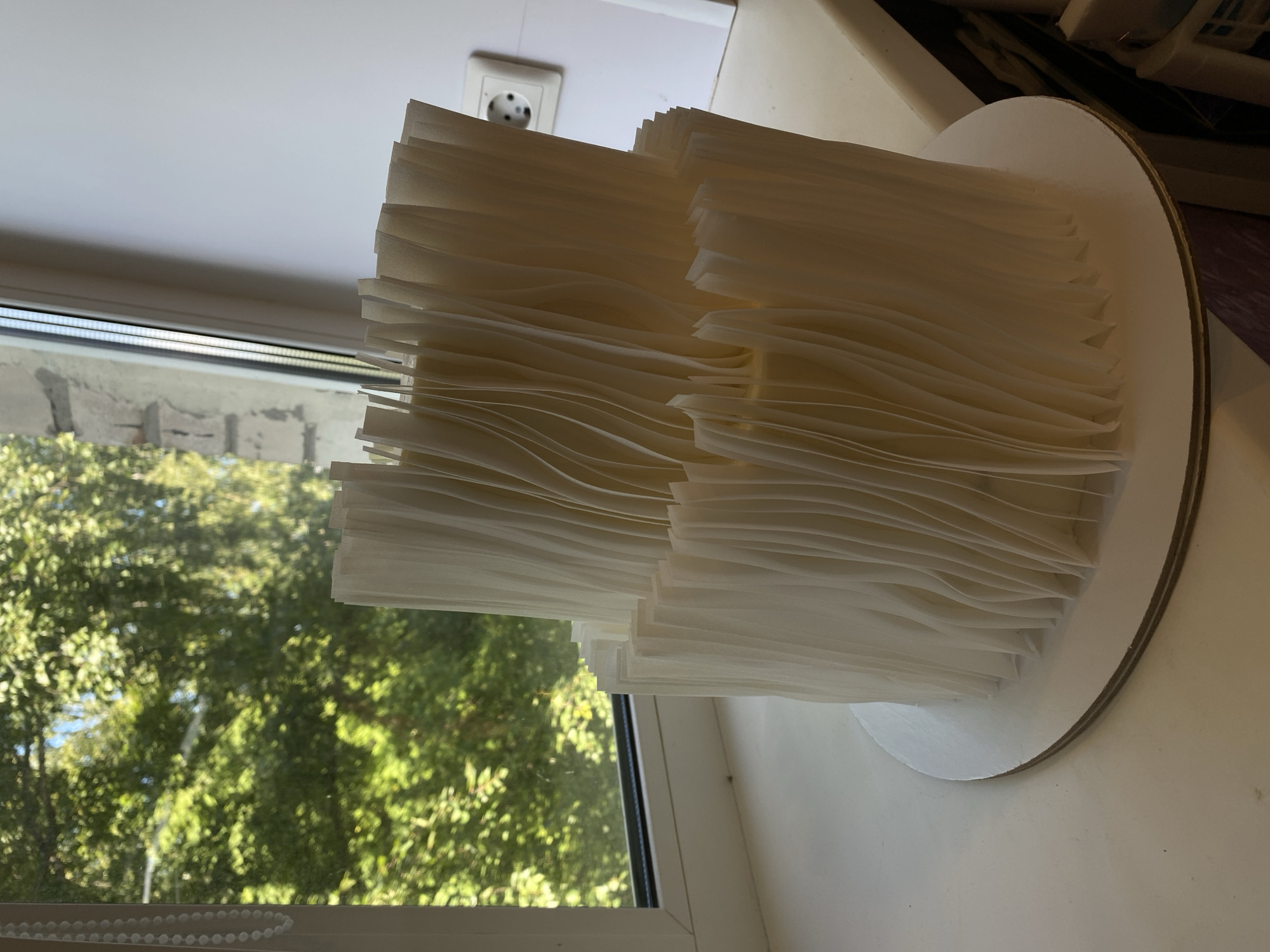 A two-tiered cake, decorated with a fan-like pattern of cream-colored edible paper, stands on a white cake board. The cake is positioned on a white windowsill, with a window to its left overlooking lush green trees and foliage. The window frame is white, and a section of a white wall with an electrical outlet is visible to the right of the cake. The background suggests an indoor setting, possibly a home or a bakery, located in the city of Lomonosov, Russia.