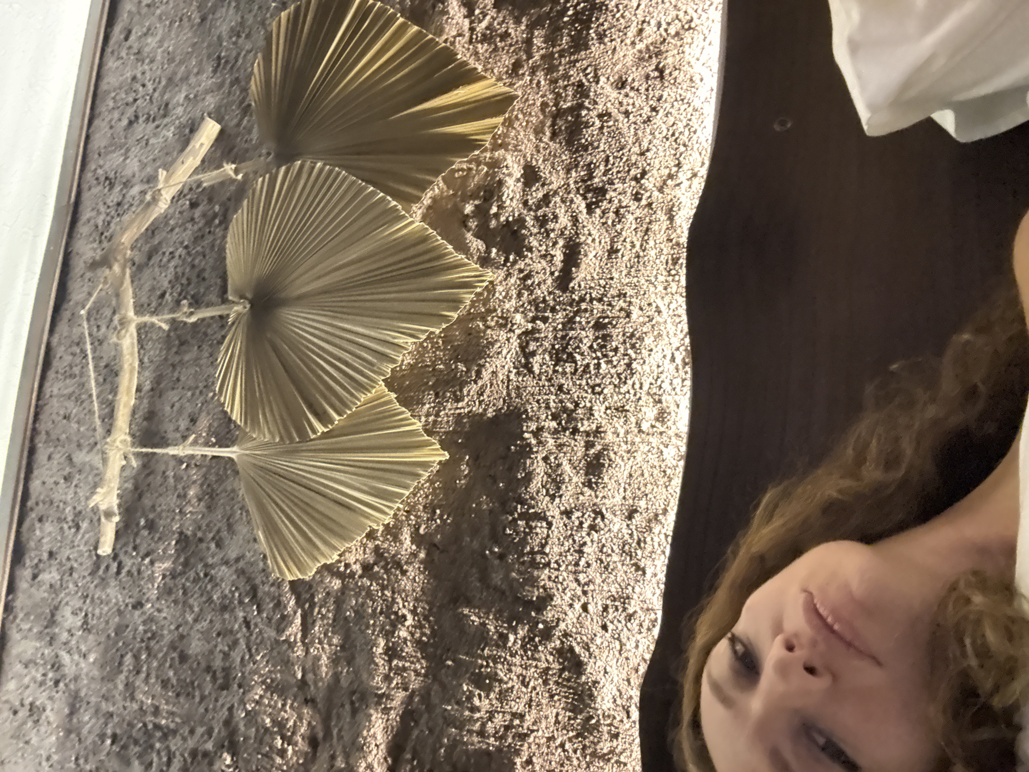 A woman is lying down, with her head and upper body visible in the bottom left of the frame. She has brown curly hair and is looking towards the camera with a neutral expression. Her eyes are dark, and she is wearing makeup. The background is a textured wall with a decorative element of three dried fan-shaped leaves hanging from a branch. The leaves are light brown and have a radiating pattern. The wall is lit from below, creating shadows and highlighting its rough texture. The overall lighting suggests an indoor setting, possibly a bedroom or a lounge area. There is a hint of a white pillow or duvet in the bottom right corner. There is no indication of the location being in Nam Hoa Lư Ward, Vietnam.