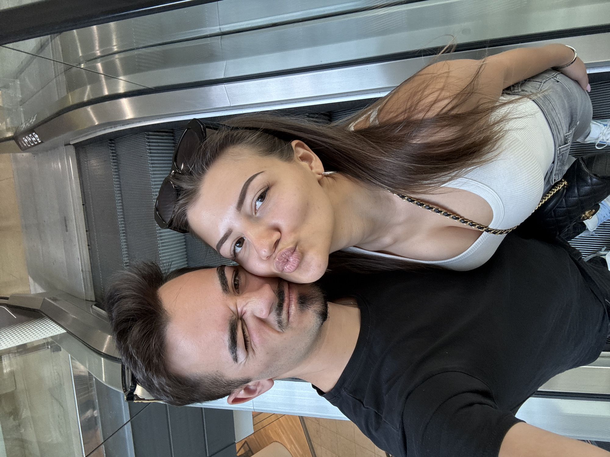 A selfie of a young Caucasian woman and man on an escalator, oriented sideways, is recorded from an overhead angle. The woman, positioned above the man, has long brown hair, and is wearing a white tank top, grey denim jeans, and sunglasses perched atop her head. She is pursing her lips towards the man’s face. The man, below, wears a black t-shirt. His face shows a comical squint. The background consists of the escalator's metallic treads and side panels. The location is presumed to be a public place within the city of Sofia, Bulgaria.