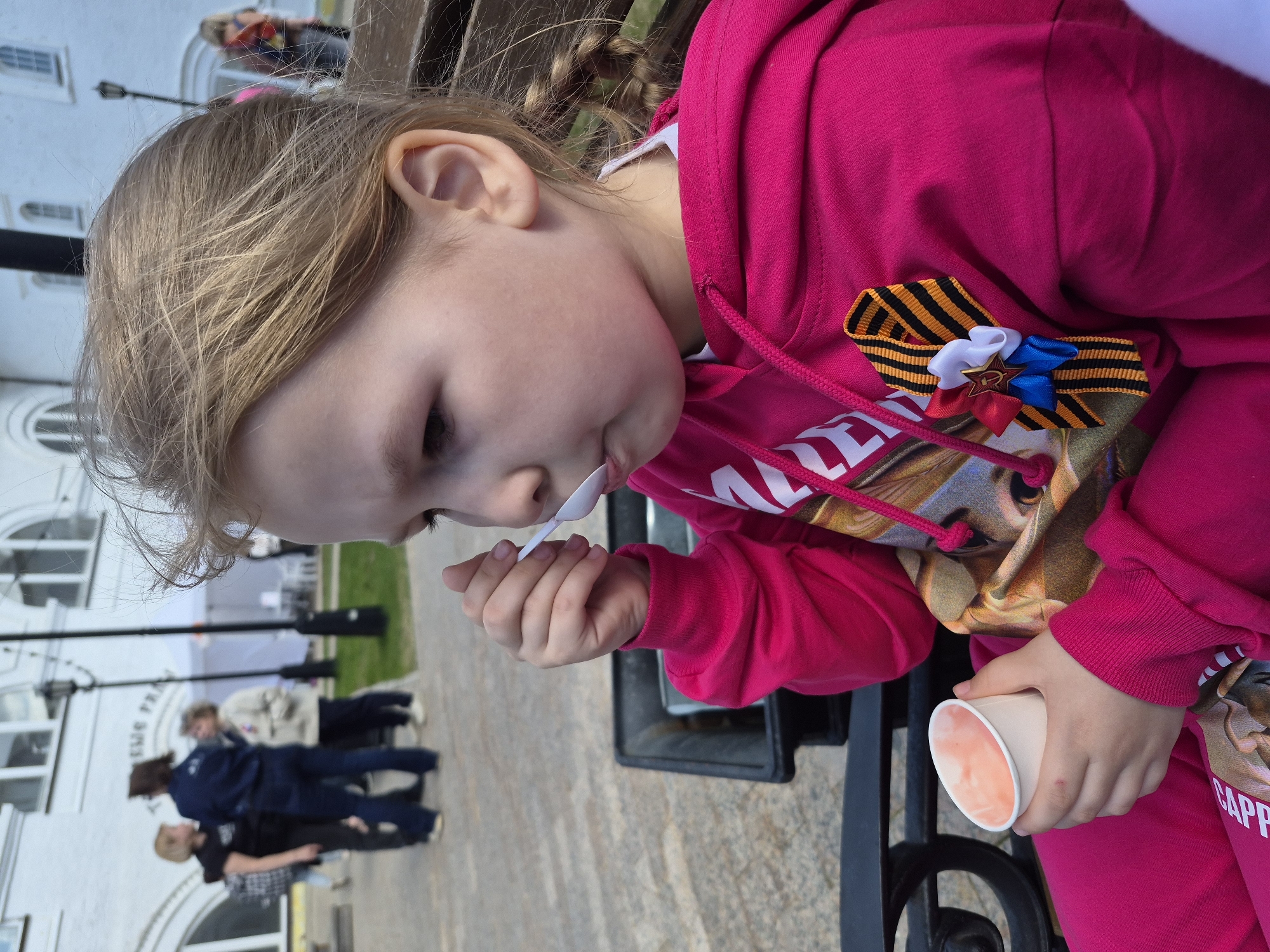 A young girl is eating from a white cup with a small white spoon while sitting on a black metal bench in Tobolsk, Russia. She is wearing a bright pink hooded sweatshirt with a printed design and matching pink pants. A Saint George ribbon and a decorative tricolor ribbon with a star are pinned to her sweatshirt. Her blonde hair is partially braided.
In the background, pedestrians are visible on a paved area, and a light-colored building with arched windows is also visible. A vintage lamp post stands nearby. The image is rotated counter-clockwise, resulting in a tilted perspective.